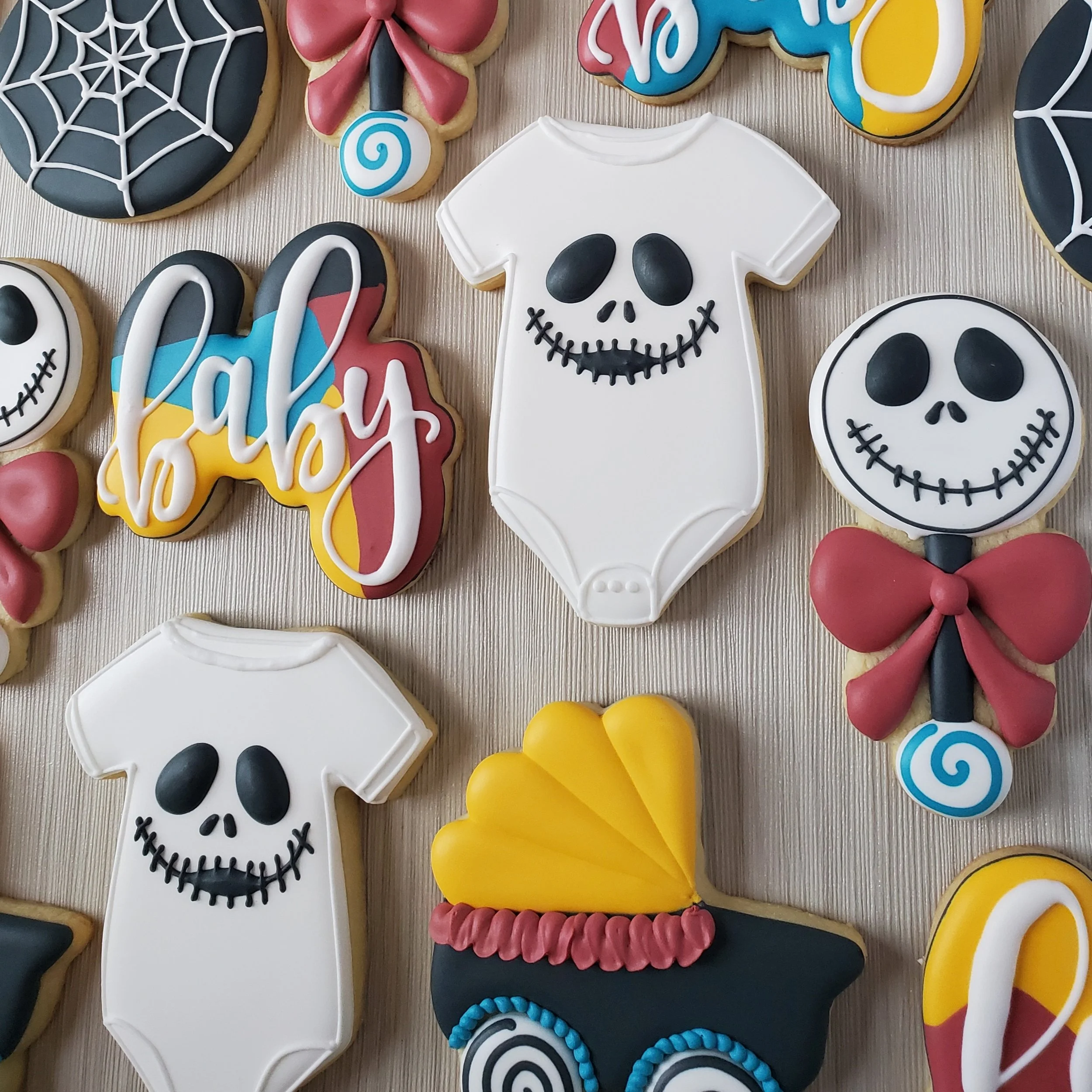 Decorated Halloween cookies including skull faces on onesie-shaped cookies, a 'baby' sign, a black cauldron with a yellow lid, and various colorful themed designs.