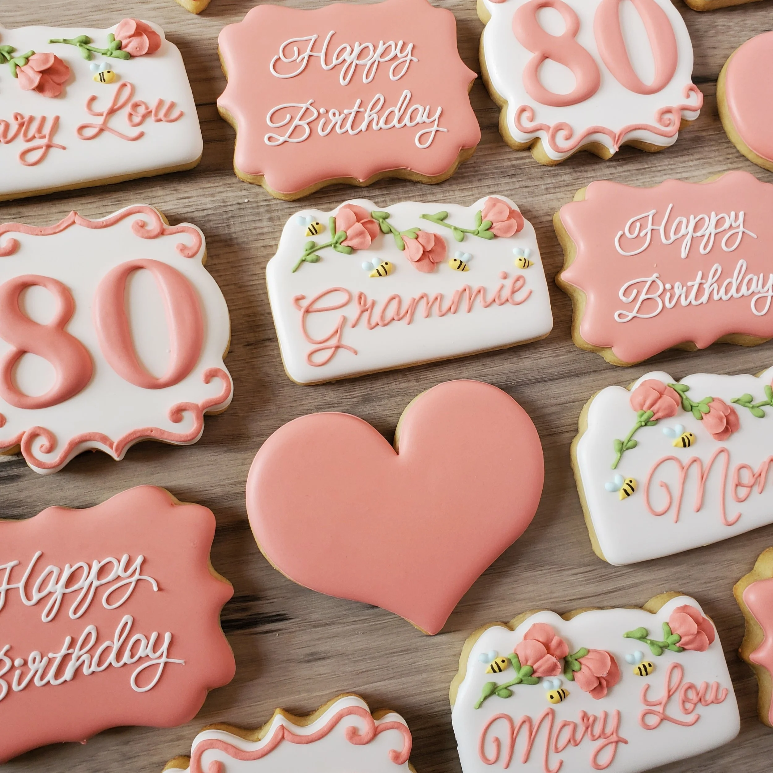 Decorative birthday cookies with pink and white icing, featuring wishes like 'Happy Birthday' and 'Mary Lou,' the number 80, a pink heart, and floral and bee embellishments.