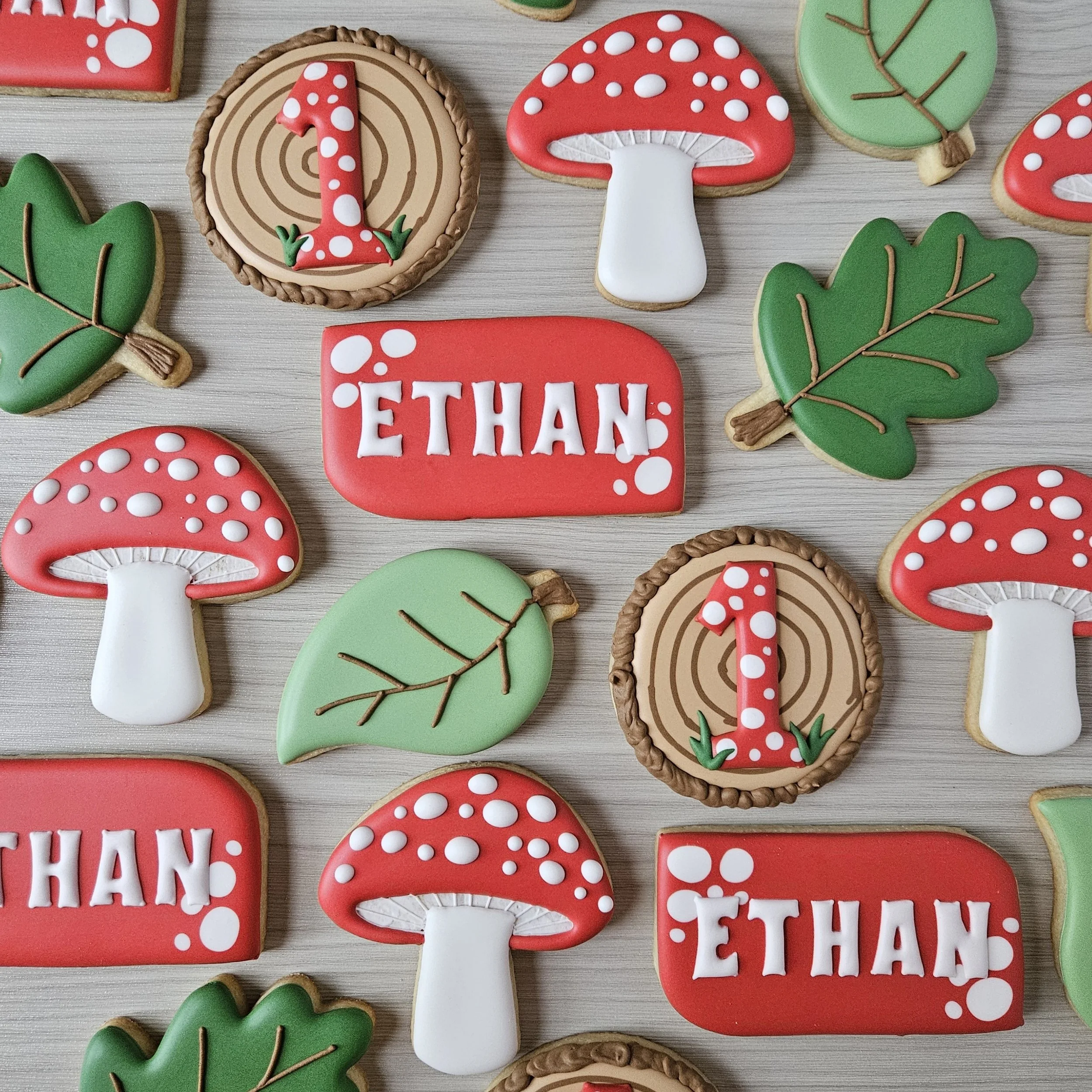 Assorted decorated cookies with mushroom, leaf, and name Ethan with a mushroom design, arranged on a light-colored surface.