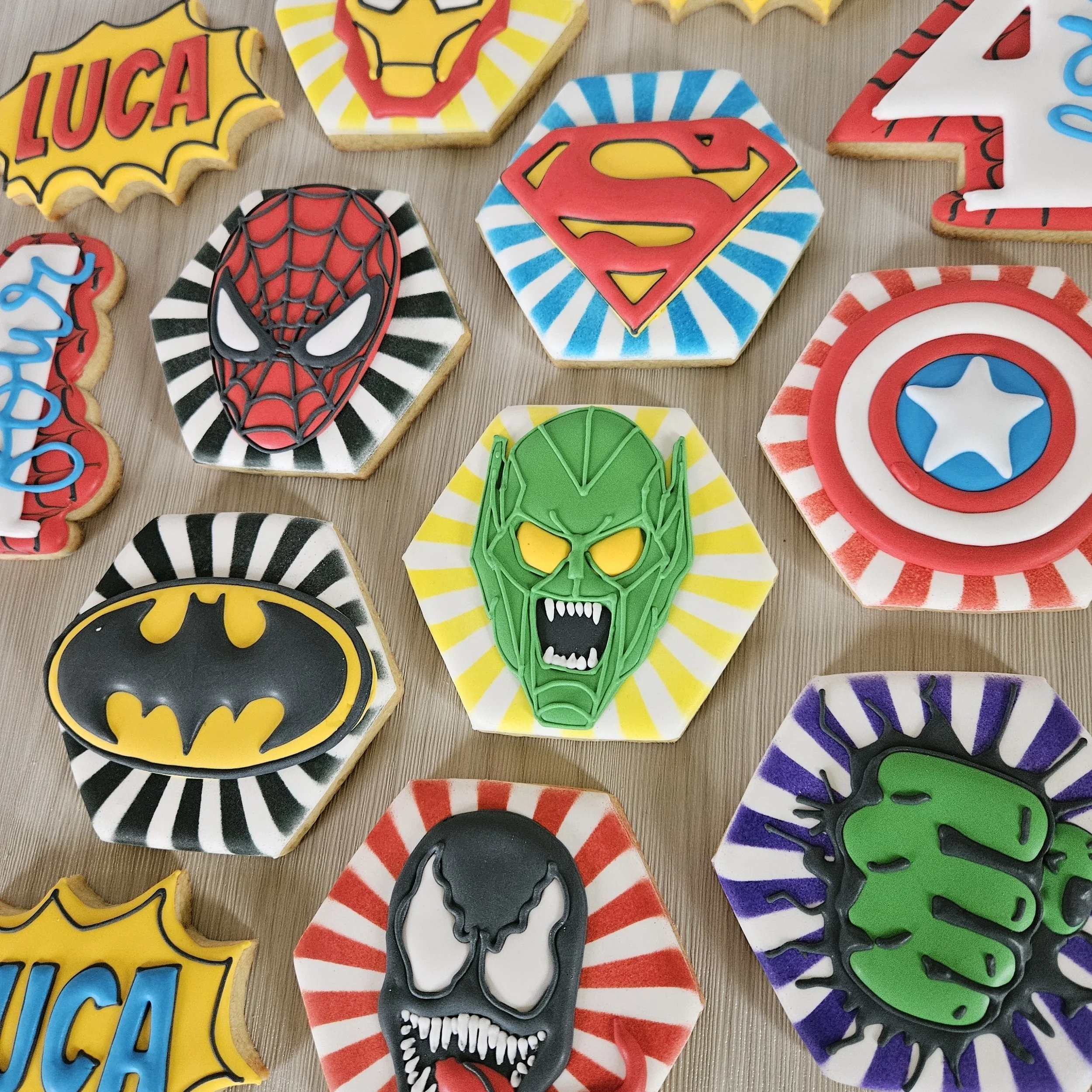 Collection of superhero-themed cookies featuring logos and characters from Marvel and DC Comics, including Spider-Man, Superman, Batman, The Hulk, Thor, Captain America, and Venom.