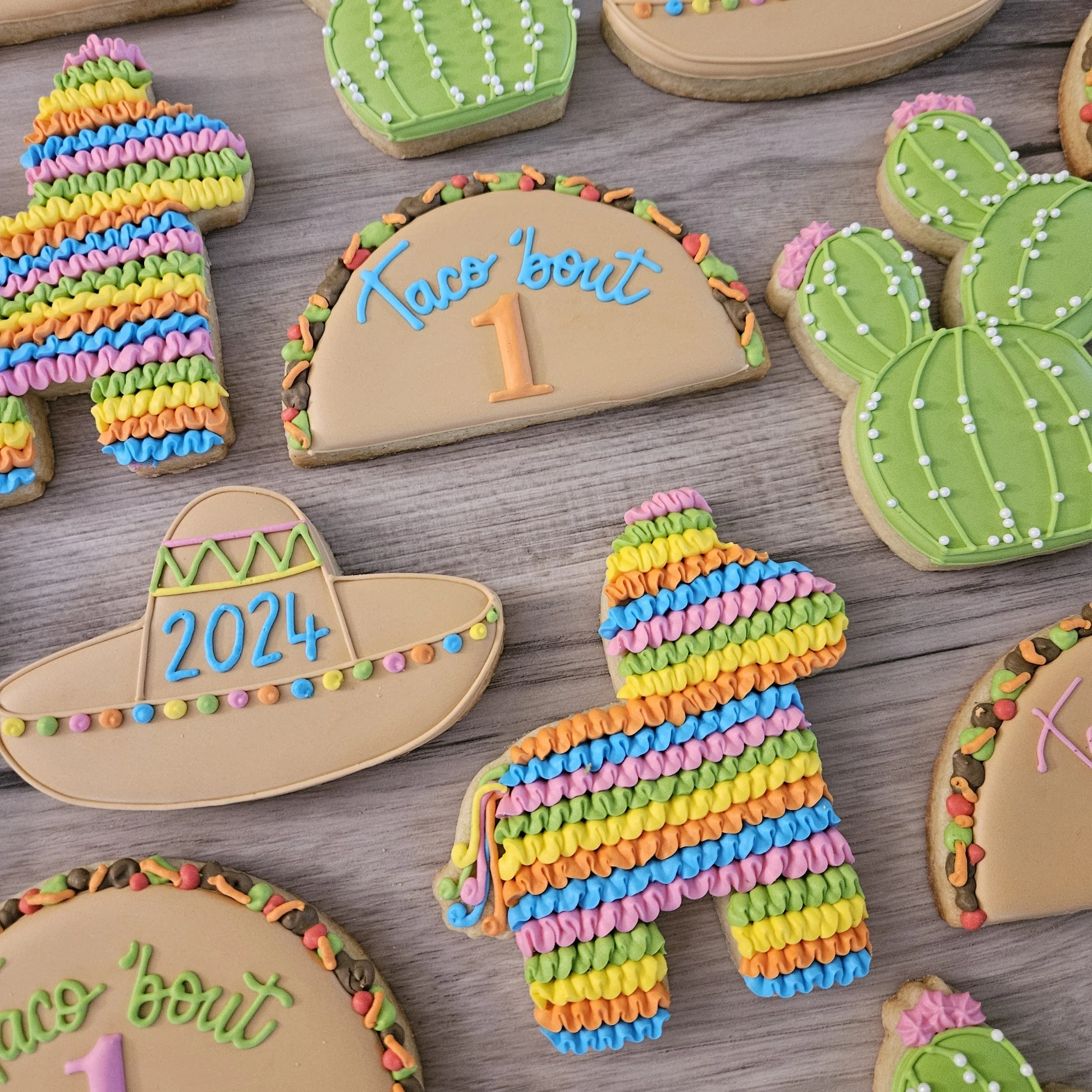 Various decorated cookies on a wooden surface, including a rainbow striped llama, cacti, a Mexican sombrero with the year 2024, a rainbow trail, and a half-circle cookie with the message "Taco 'bout 1" for a first birthday celebration.