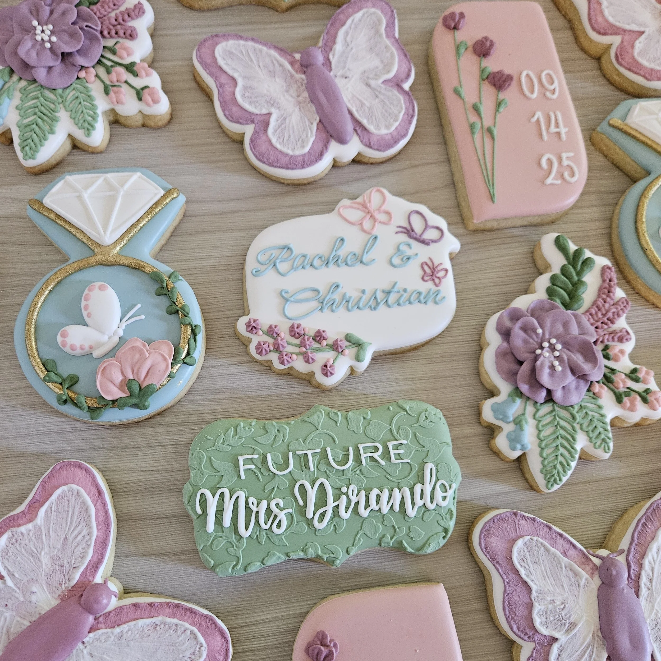 Decorative cookies with floral, butterfly, and message designs, including one shaped like a diamond ring and a wedding plaque with the names Rachel and Christian and wedding date 09/14/25, arranged on a wooden surface.