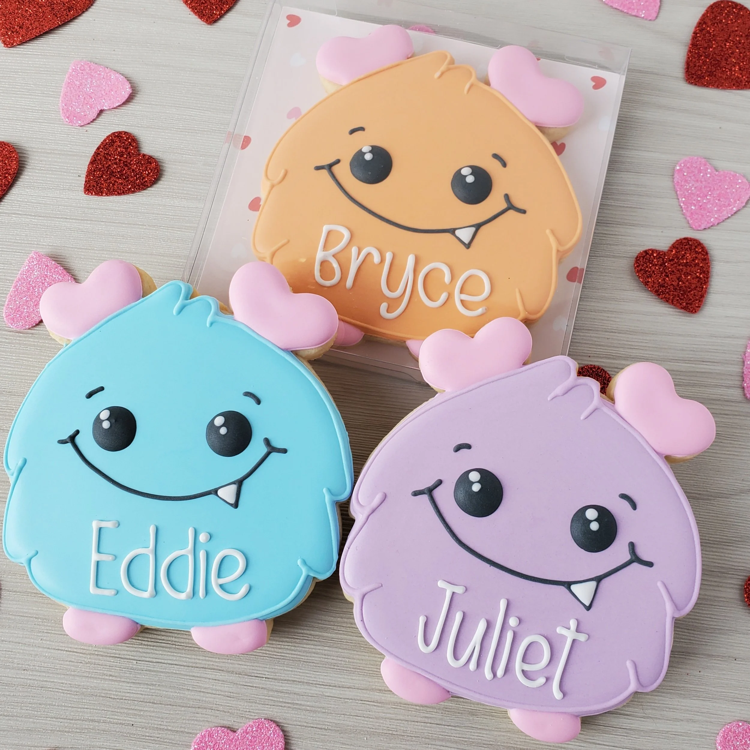 Decorated cookies shaped like cute monsters with pink ears, each with a different name: Bruce, Eddie, and Juliet. The cookies are pastel-colored in peach, blue, and purple, surrounded by pink and red glittery hearts on a light-colored wooden surface.
