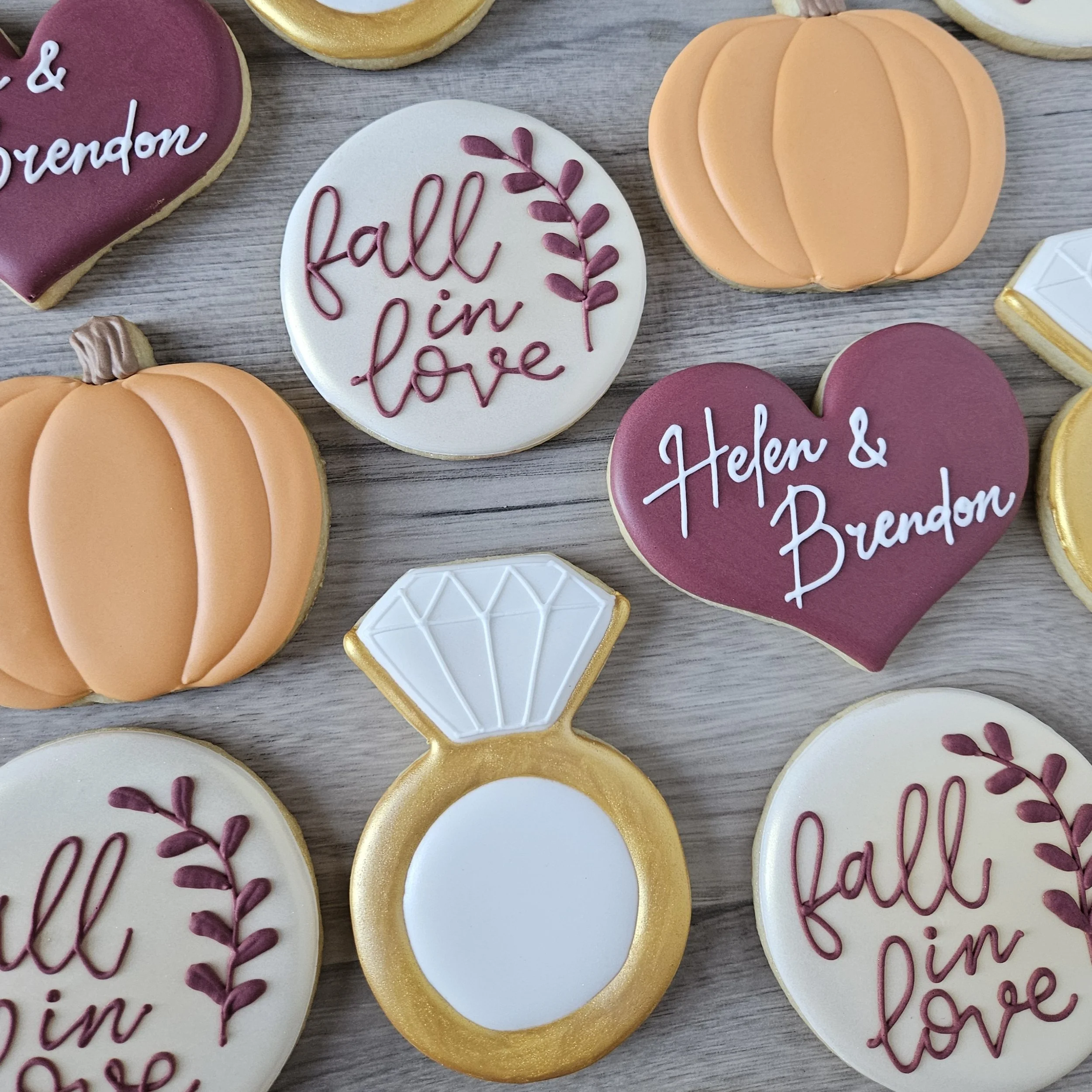 Decorative cookies with fall and love themes, including pumpkins, a diamond ring, and hearts with inscriptions like "fall in love" and "Helen & Brandon" on a wooden surface.