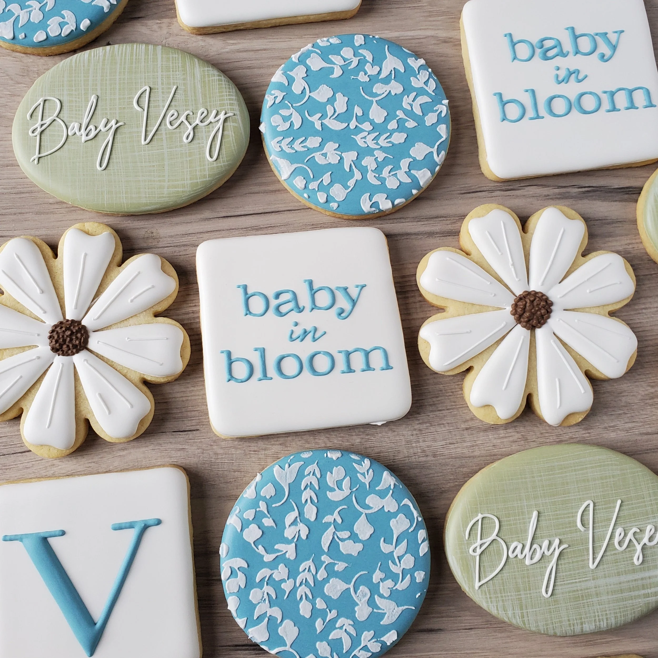 Decorative cookies with floral and text designs, in blue, white, and green, with phrases like 'baby in bloom' and 'Baby Vesey' and floral patterns, arranged on a wooden surface.