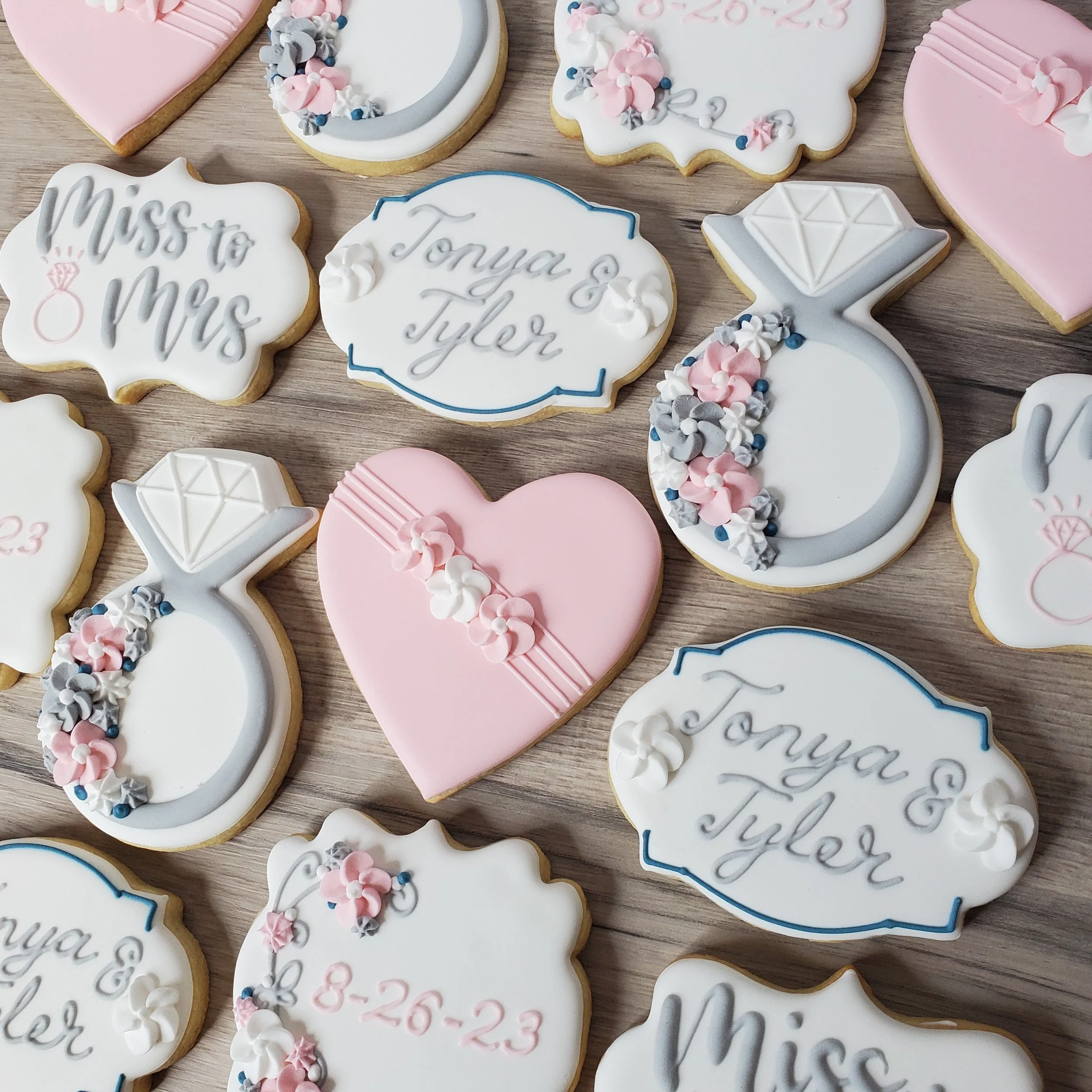 Decorated cookies with wedding and engagement themes, including a pink heart, a diamond ring, and signs with names 'Tonya & Tyler' and 'Miss to Mrs', and wedding date '8-26-23', laid out on a wooden surface.