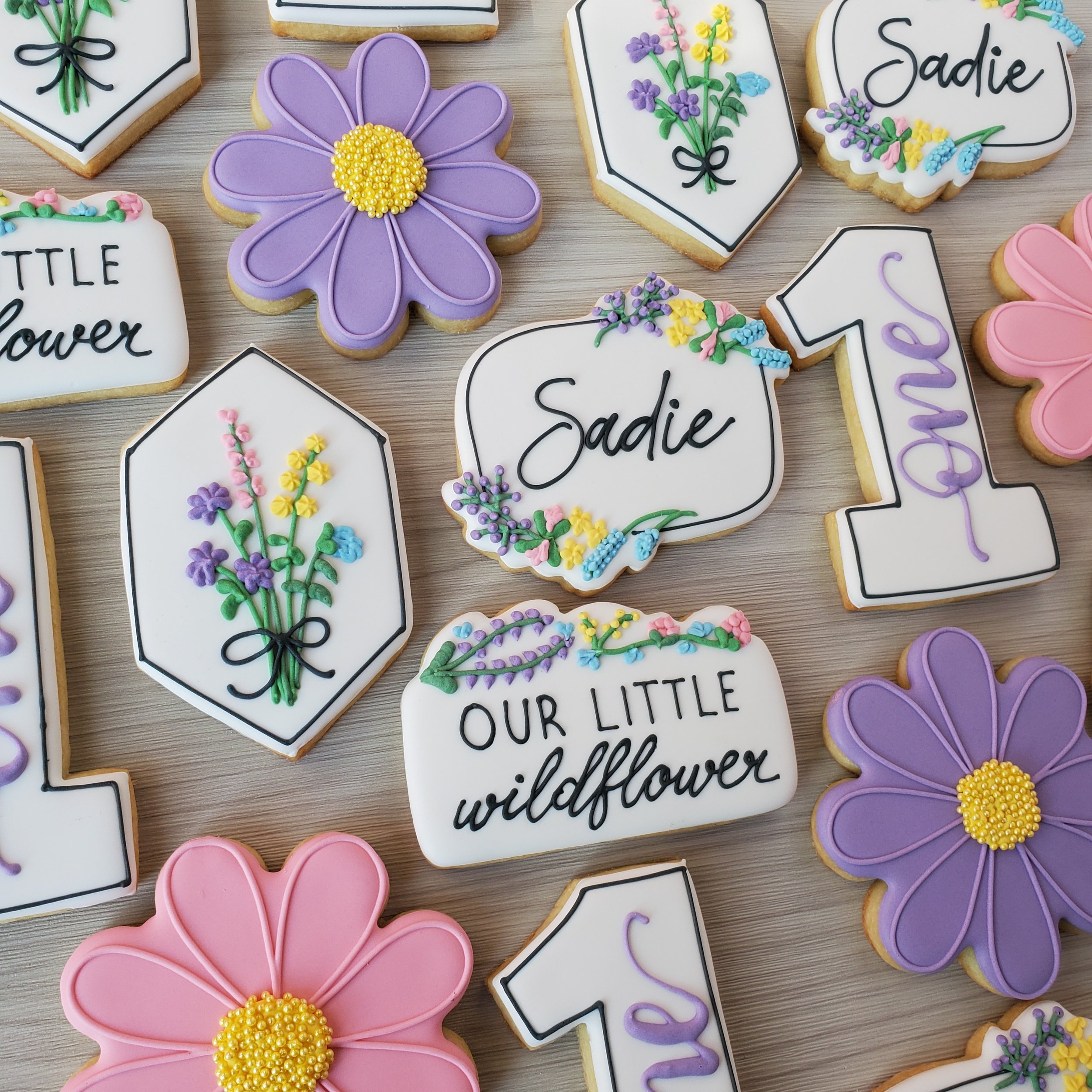 Decorative cookies with flowers and words, including purple flowers, pink flowers, yellow, purple, and blue flower designs, and messages like 'Sadie' and 'Our little wildflower' on white backgrounds.