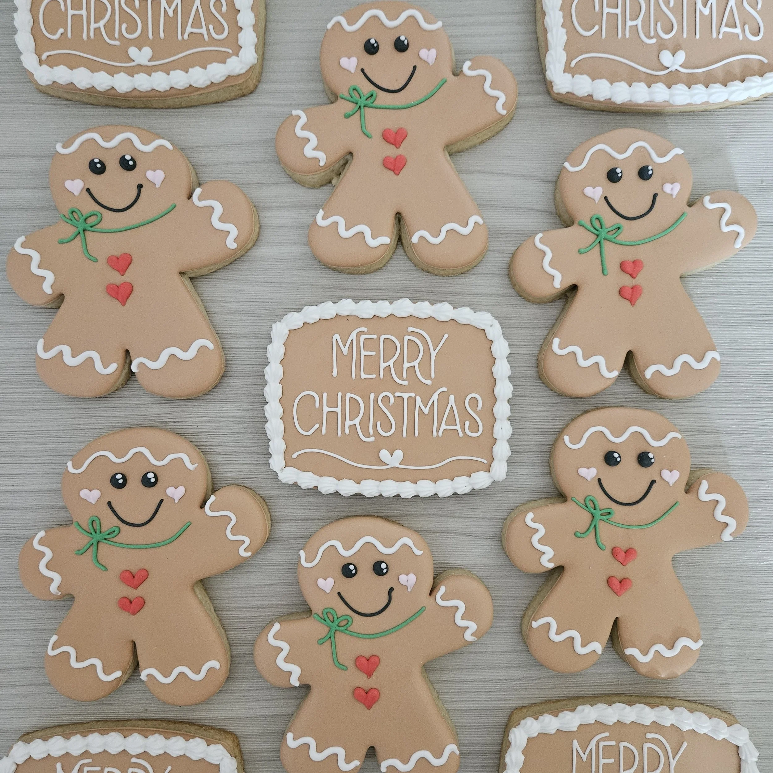 Christmas-themed sugar cookies decorated as gingerbread men with smiling faces, green bows, red hearts, and white icing accents, arranged around a 'Merry Christmas' message written on a square cookie.