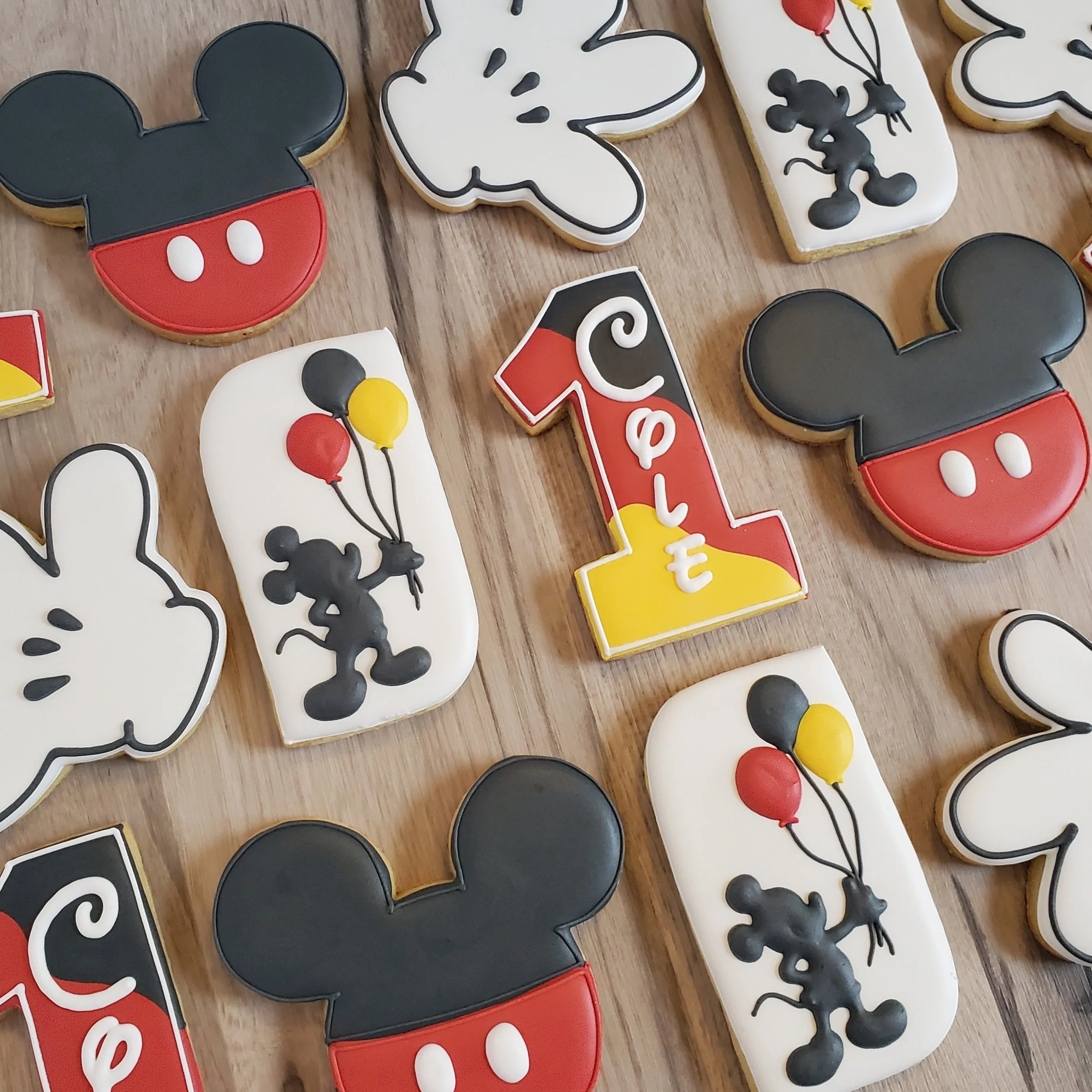 Assorted Mickey Mouse themed cookies decorated with Mickey Mouse heads, butterflies, balloons, and the number 1 with a birthday message on a wooden surface.