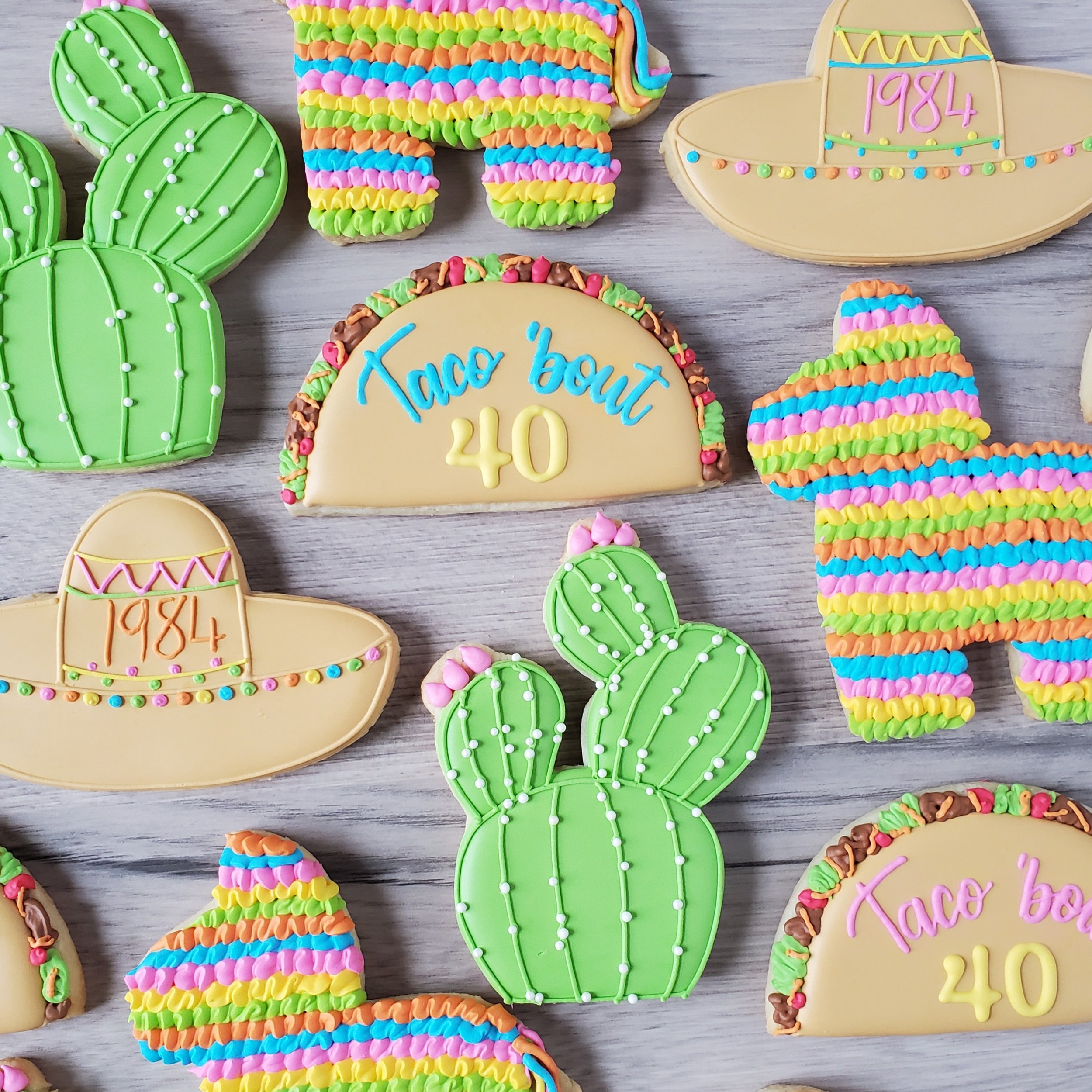 Colorful decorated cookies shaped like cacti, sombreros, and a rainbow pinata, with one cookie reading "Taco bout 40" in blue and yellow icing, arranged on a light wooden surface.