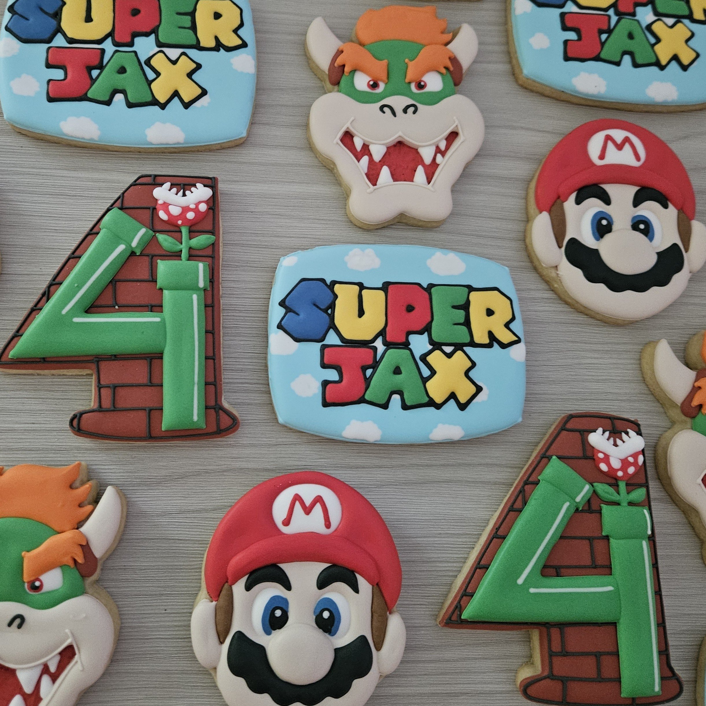 Decorated cookies themed around Super Mario. Some cookies depict the words 'Super Jax' with clouds, others show a cartoonish Luigi and Mario faces, and a couple feature large colorful number 4s with brick patterns and a green pipe with a piranha plan