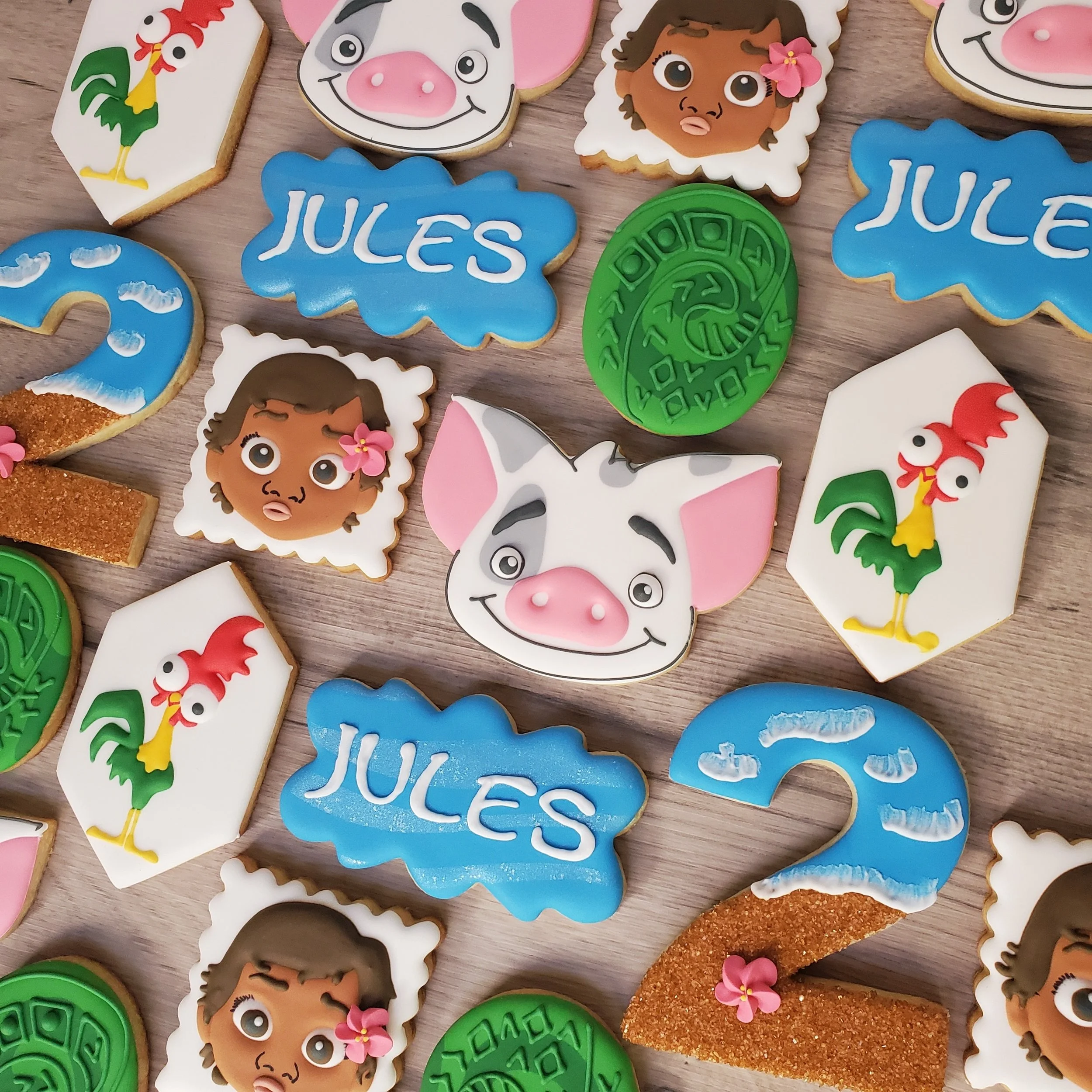 Decorative cookies with characters from Moana, including baby Moana, Pua, along with colorful designs of the number 2, the word Jules, a green heart of Tefiti, and a Hei Hei, all arranged on a wooden surface.