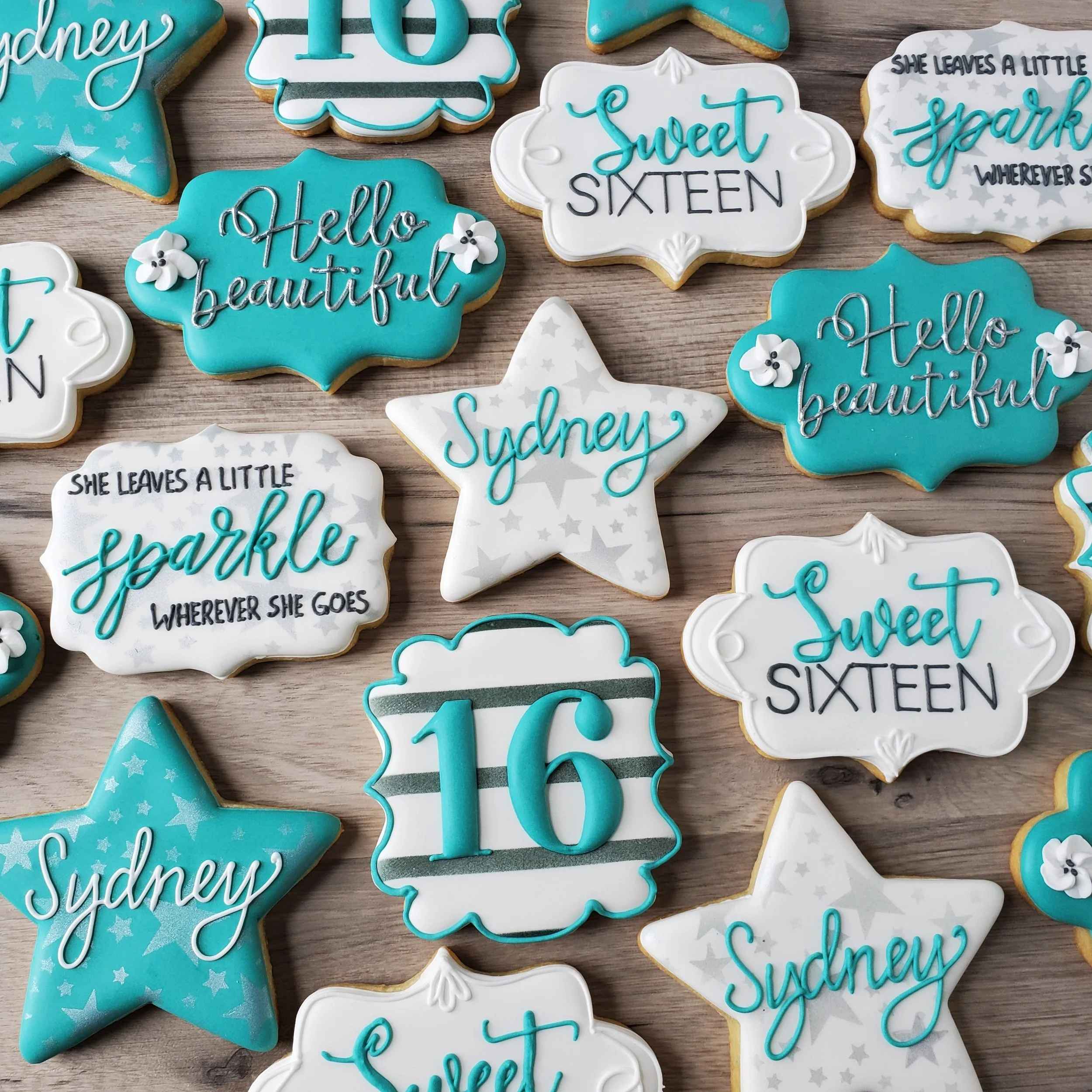 Assorted decorated cookies on a wooden surface, featuring phrases like 'Hello beautiful,' 'Sweet sixteen,' and 'She leaves a little sparkle wherever she goes,' along with star and cloud shapes, some with the name 'Sydney' and the number '16' in blue 