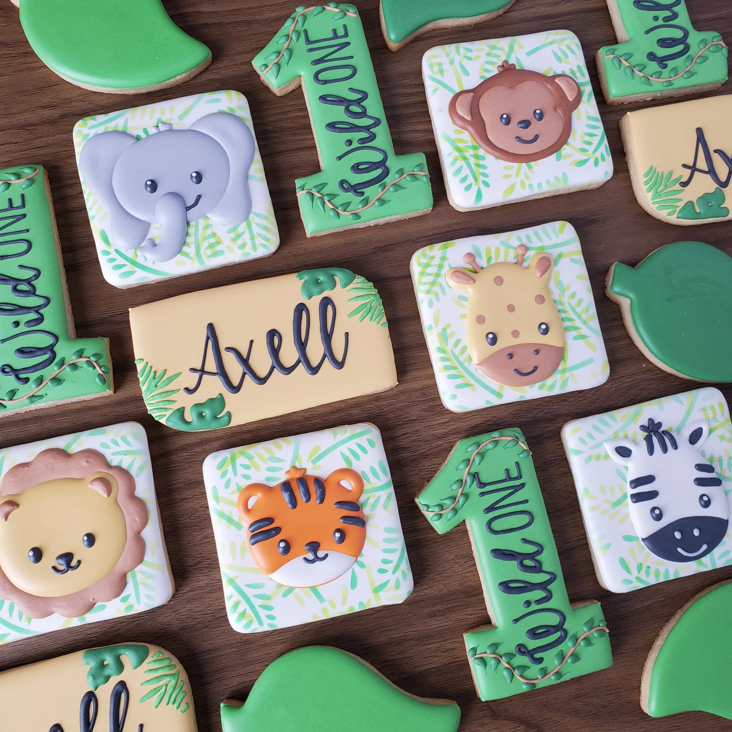 Animal-themed decorated cookies with jungle animals such as elephant, bear, giraffe, lion, tiger, and zebra, along with green leaf and plant designs and personalized names.