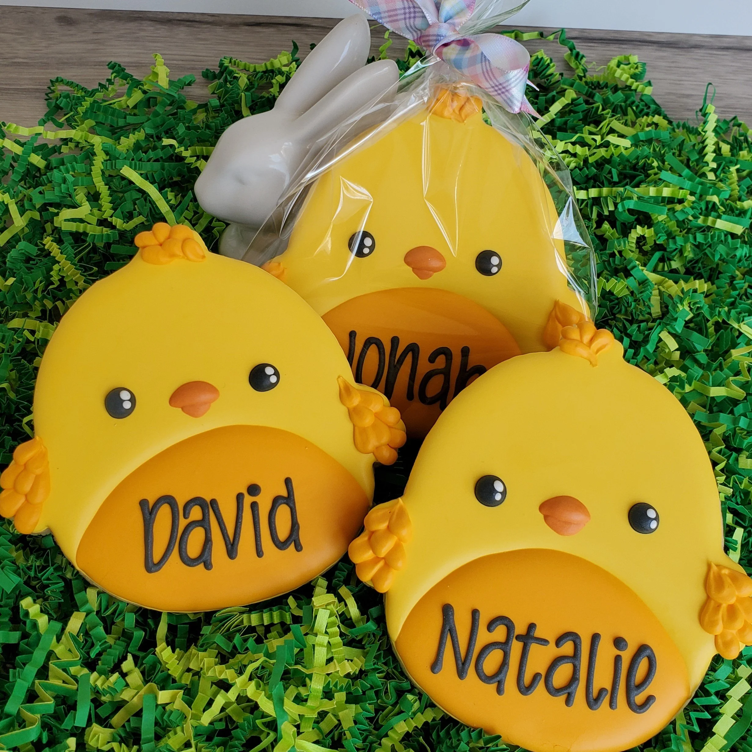 Yellow chick-shaped cookies with orange beak and chocolate eyes, decorated with orange frosting shaped like little flowers or wheat, with personalized names 'David' and 'Natalie' written on their bellies. A ceramic white bunny is also in the backgrou