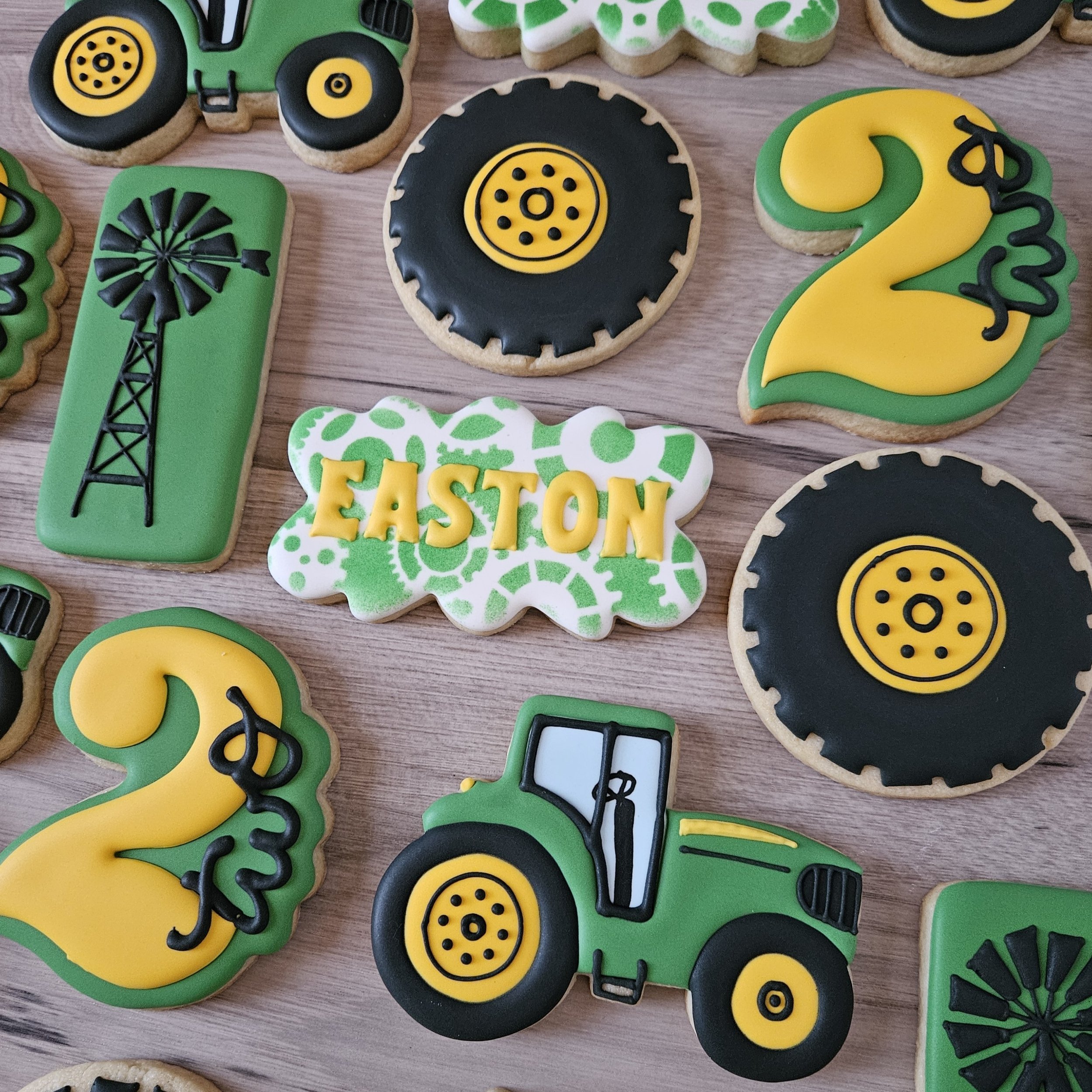 Decorated sugar cookies with a farming theme, including tractors, saw blades, the number 2, a windmill, and the name 'EASTON' on a patterned background.