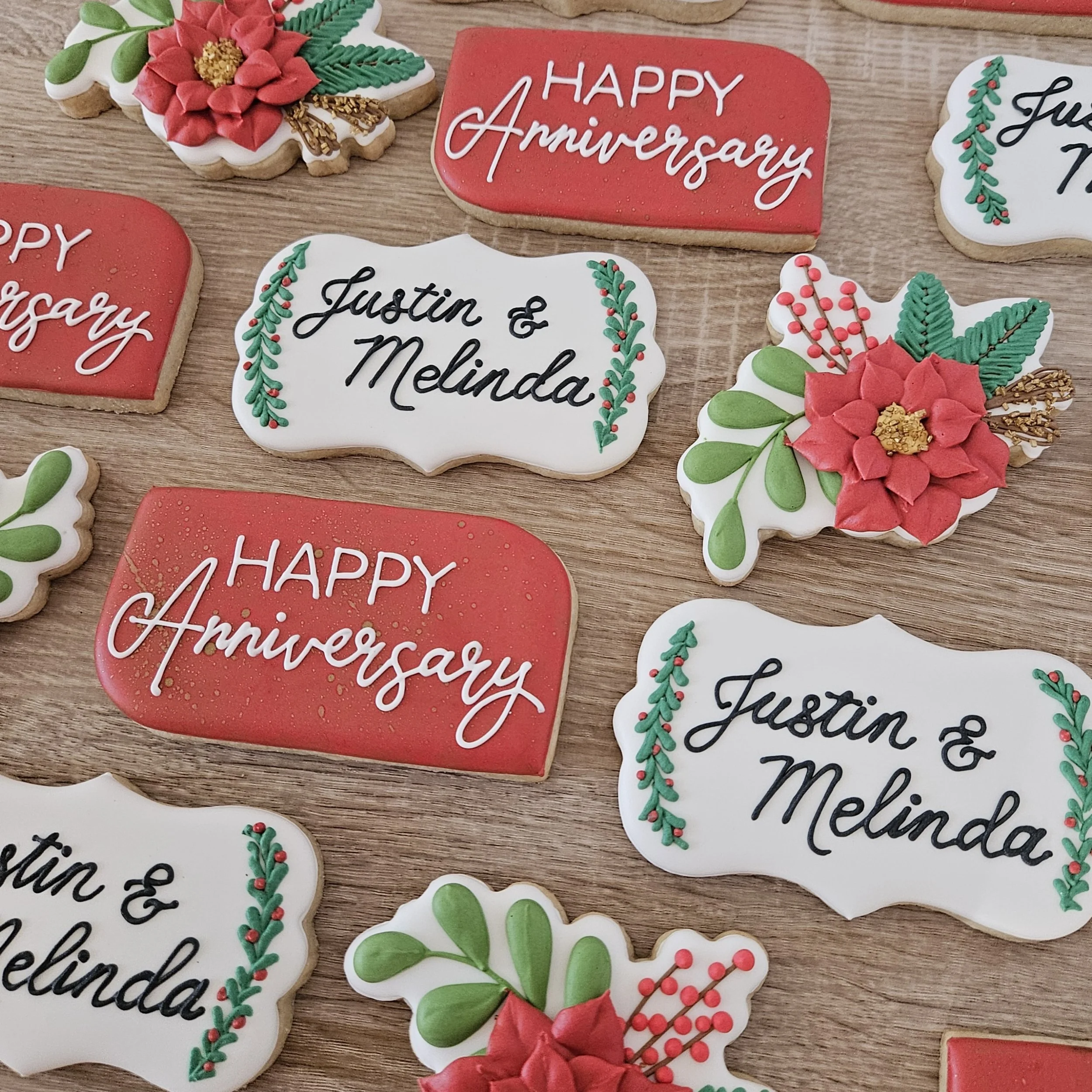 Decorative cookies with holiday themes, including red and white rectangles with messages, and poinsettia and greenery designs, arranged on a wooden surface.