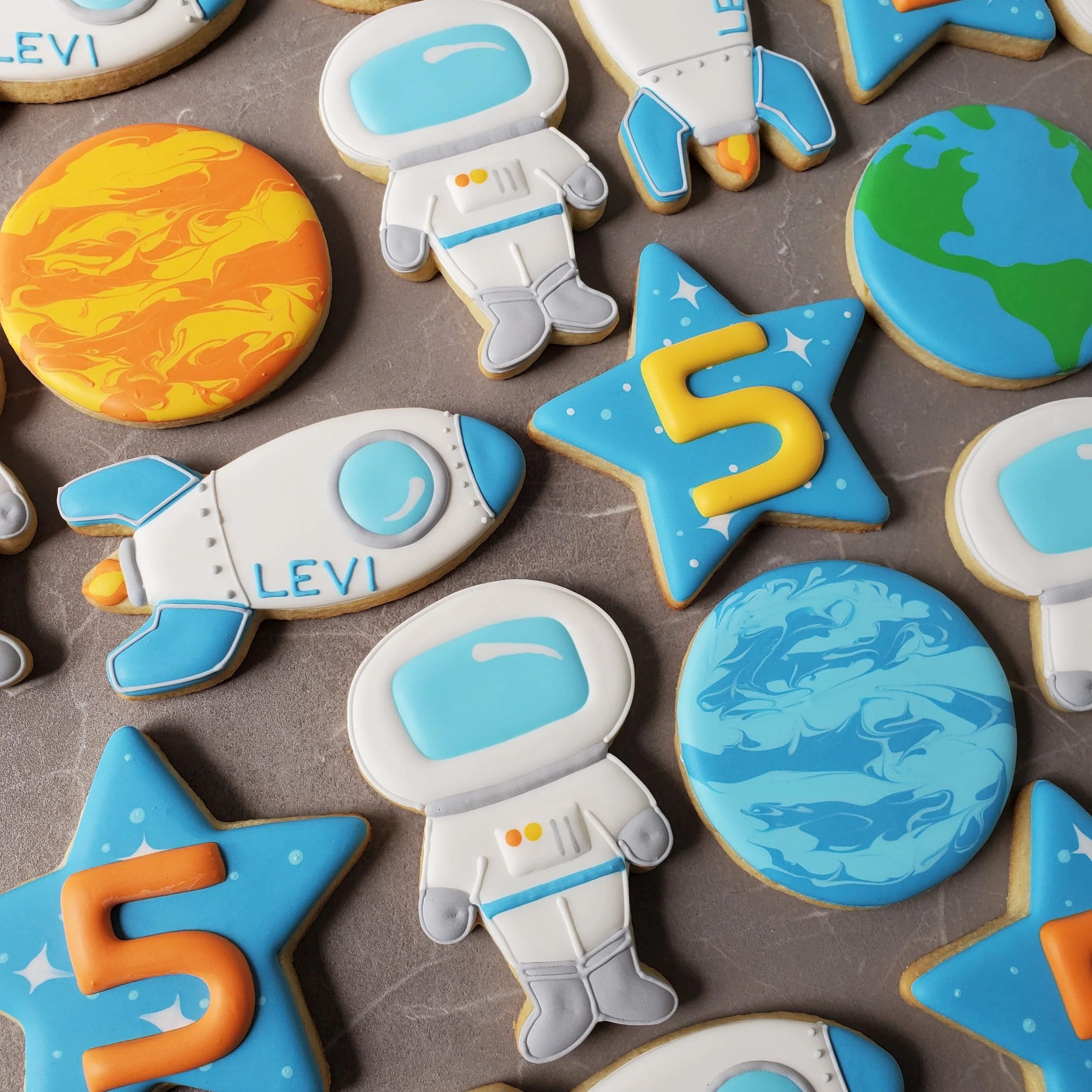 Decorated cookies with space theme, including rockets, planets, stars with the number 5, and an astronaut, on a gray surface.