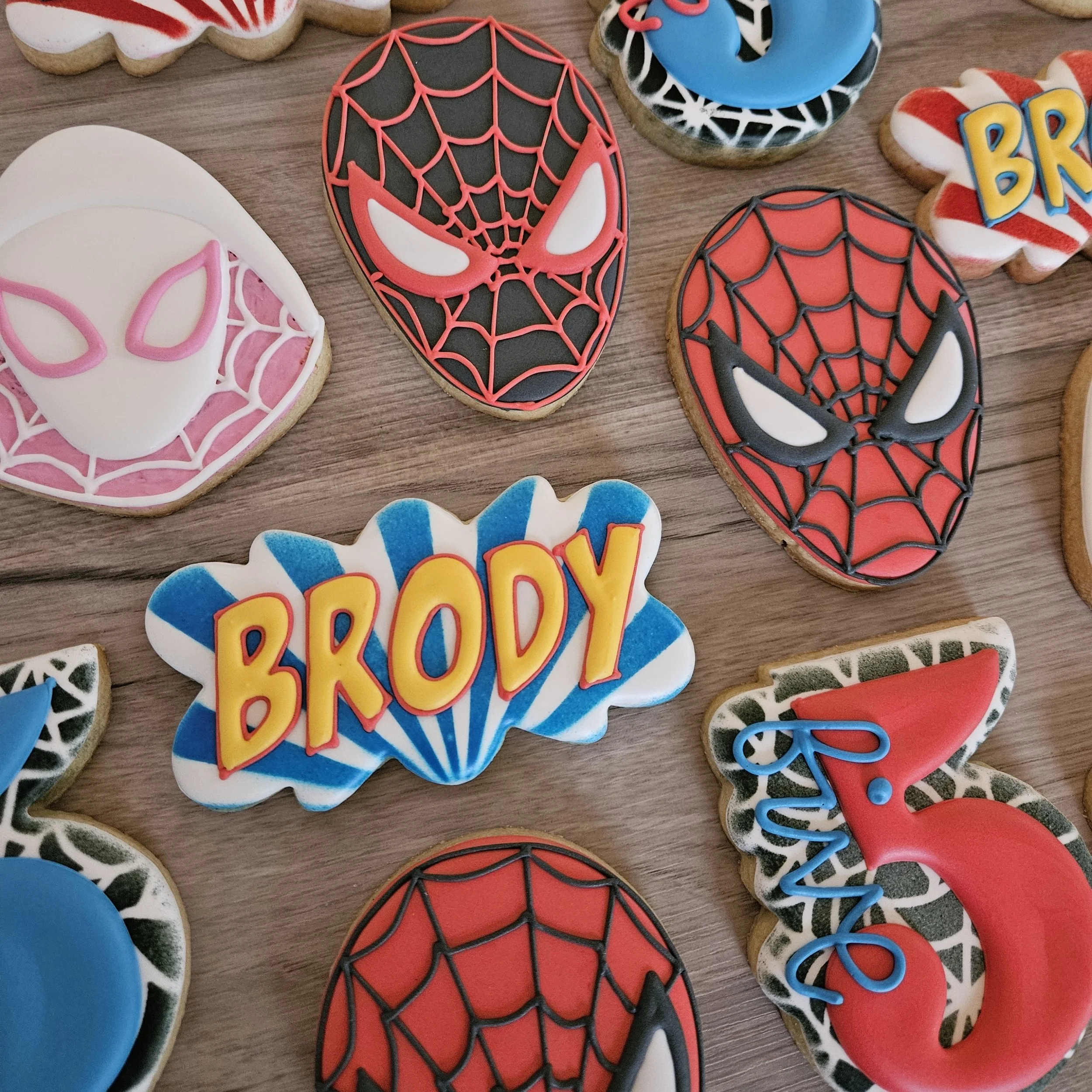 Decorated cookies with superhero and birthday themes, including Spider-Man, Spider-Woman, and a cookie with the name 'Brody' in colorful icing, displayed on a wooden surface.