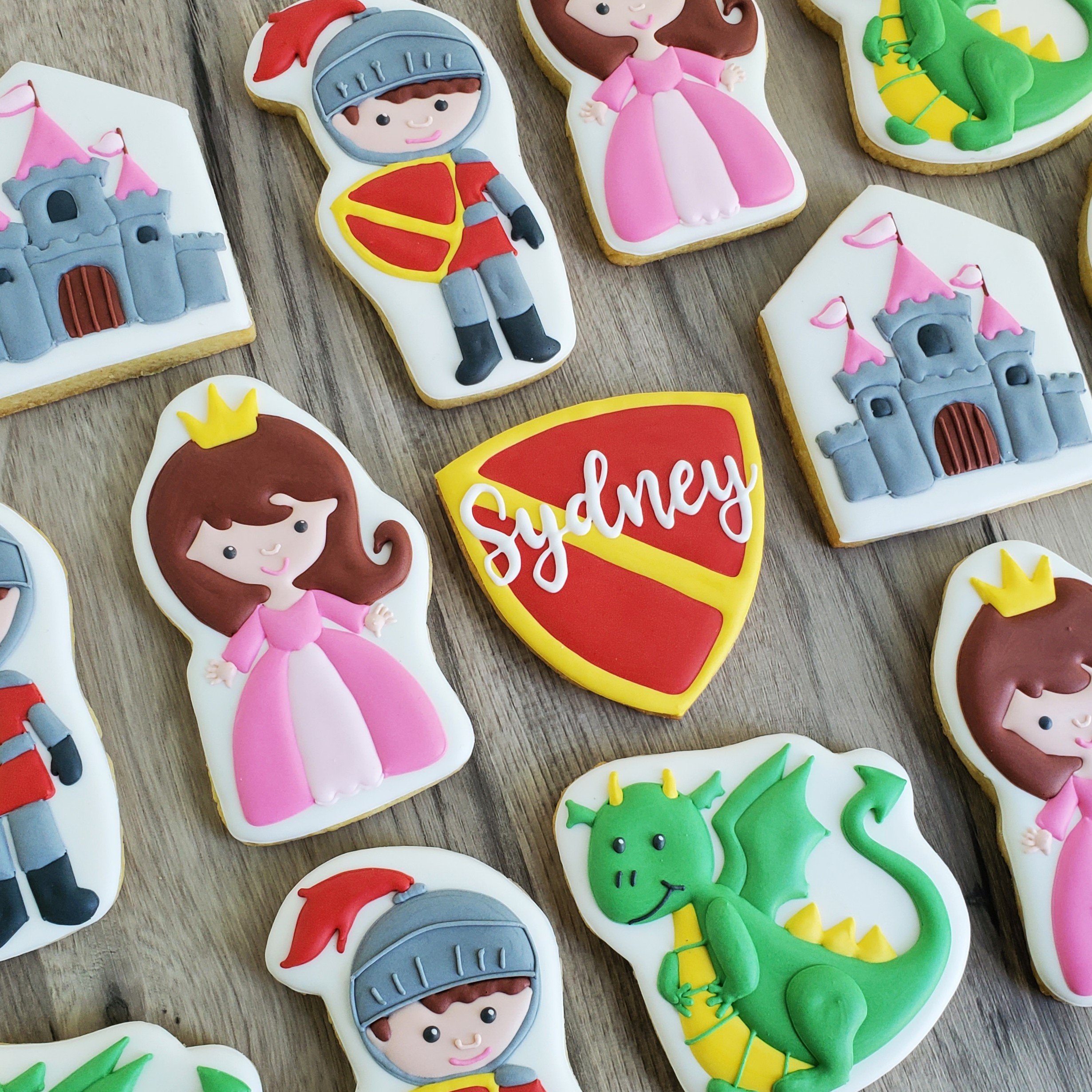 Decorated cookies featuring characters and symbols related to Disney princesses, knights, castles, and a dragon, with one cookie reading 'Sydney' in white lettering on a red and yellow shield background.