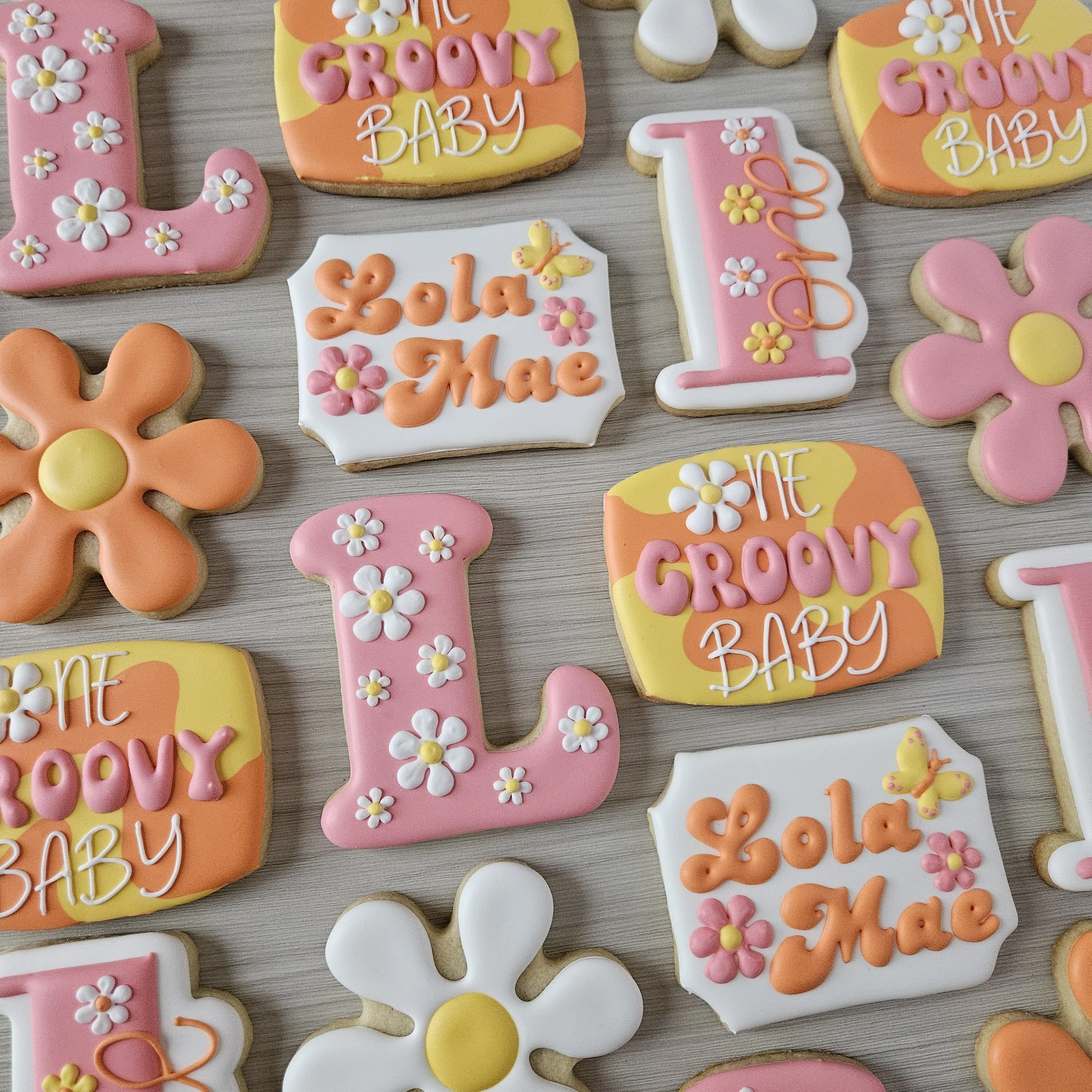 Decorated cookies for a One Groovy Baby first birthday in pinks, yellows, oranges and white. Featuring daisy flowers, a capital L with daisies, a number one, a cookie that says "Lola Mae" and another that says "One Groovy Baby"
