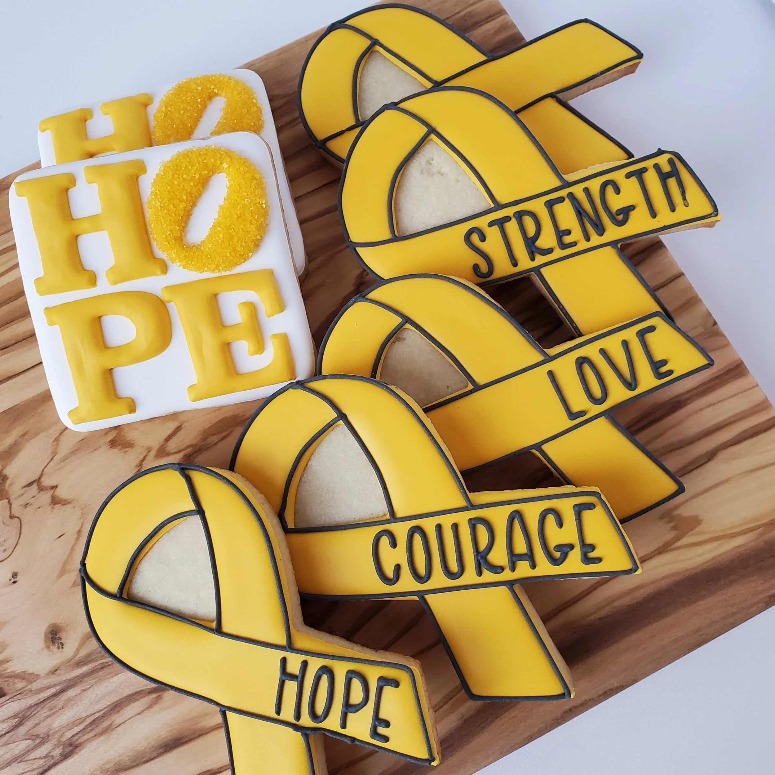 Decorative cookies on a wooden board with yellow ribbons featuring the words "Hope," "Courage," "Love," and "Strength," and a white square with yellow letters spelling "HOPE" with yellow sugar.