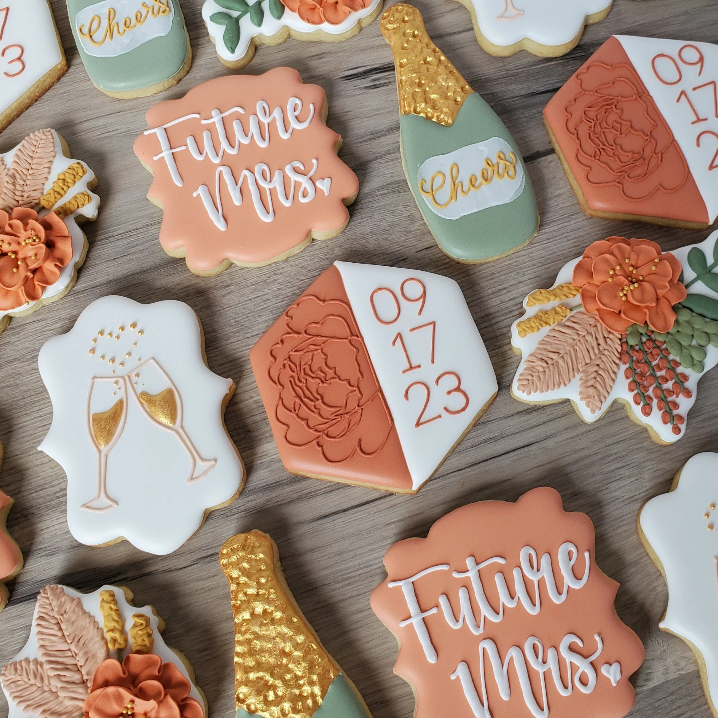 Decorative cookies with wedding and celebration themes, including a cookie labeled 'Future Mrs.', a champagne bottle, a birthday cake with dates '09 17 23', a pair of champagne glasses, and floral designs, all arranged on a wooden surface.