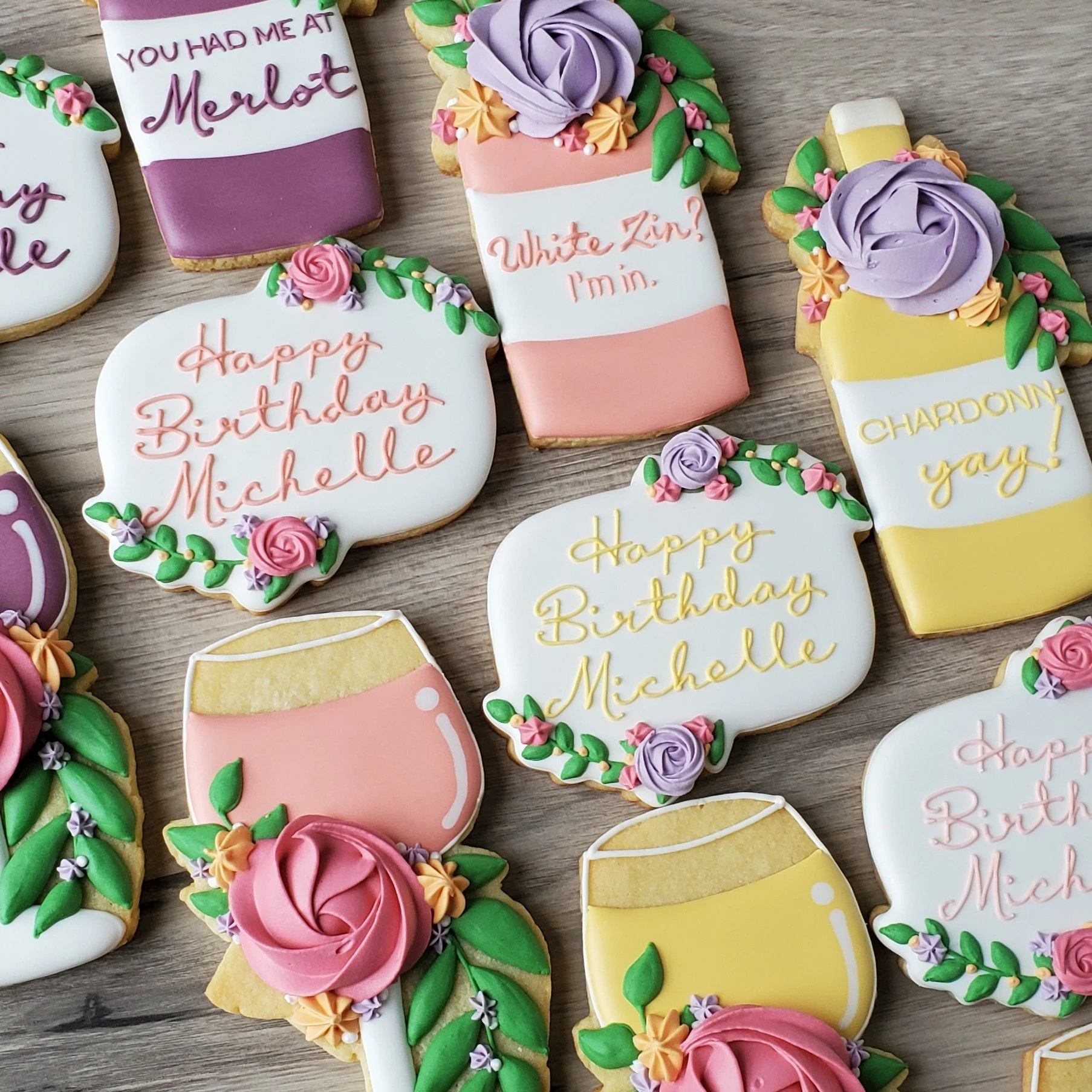 Decorated birthday cookies with floral and champagne themes, featuring messages like 'Happy Birthday Michelle' and 'White Zinf? I'm in,' on a wooden surface.