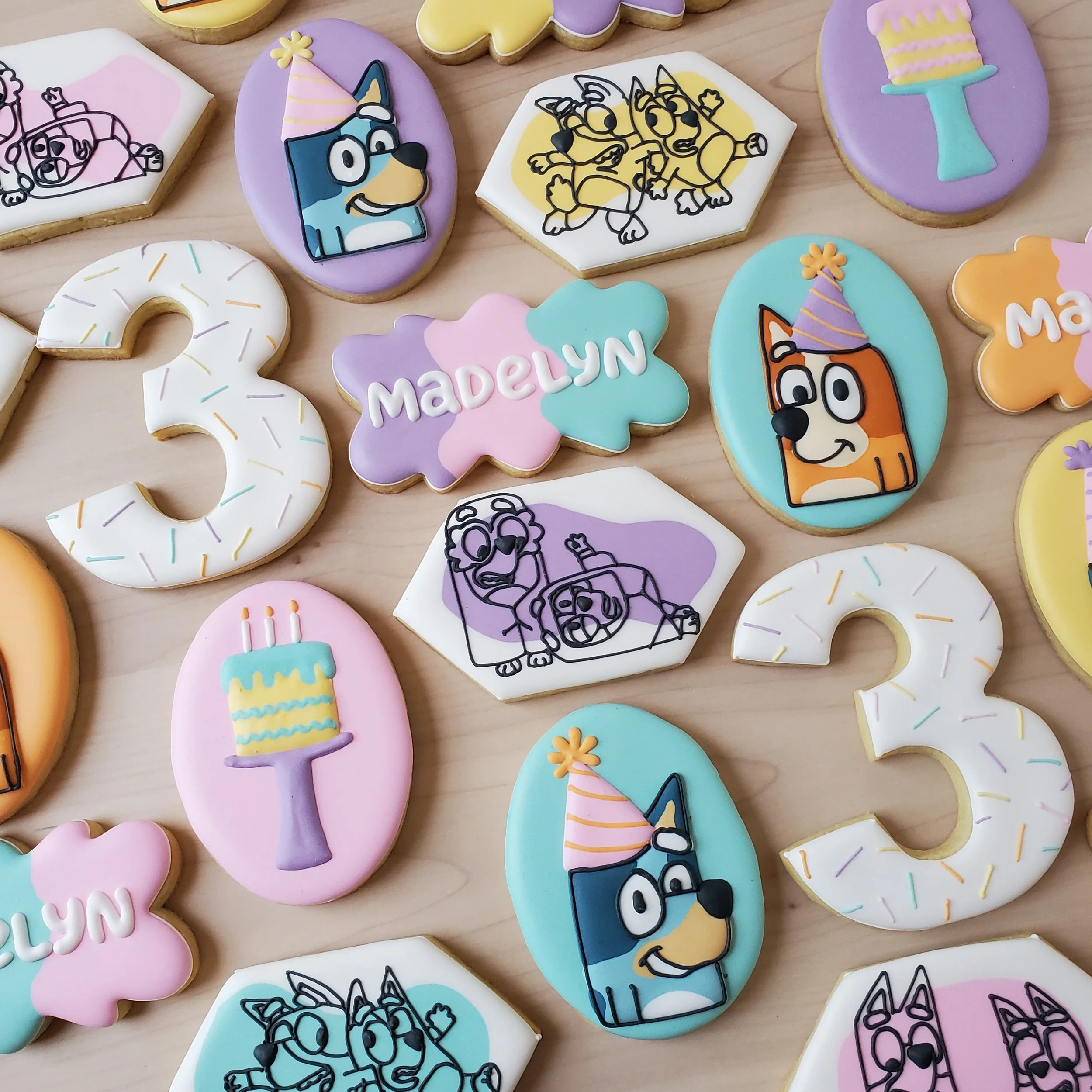 Decorated cookies with birthday themes, including a large number 3, a cake, cartoon dogs with party hats, and a cookie with the name Madelyn, on a wooden surface.