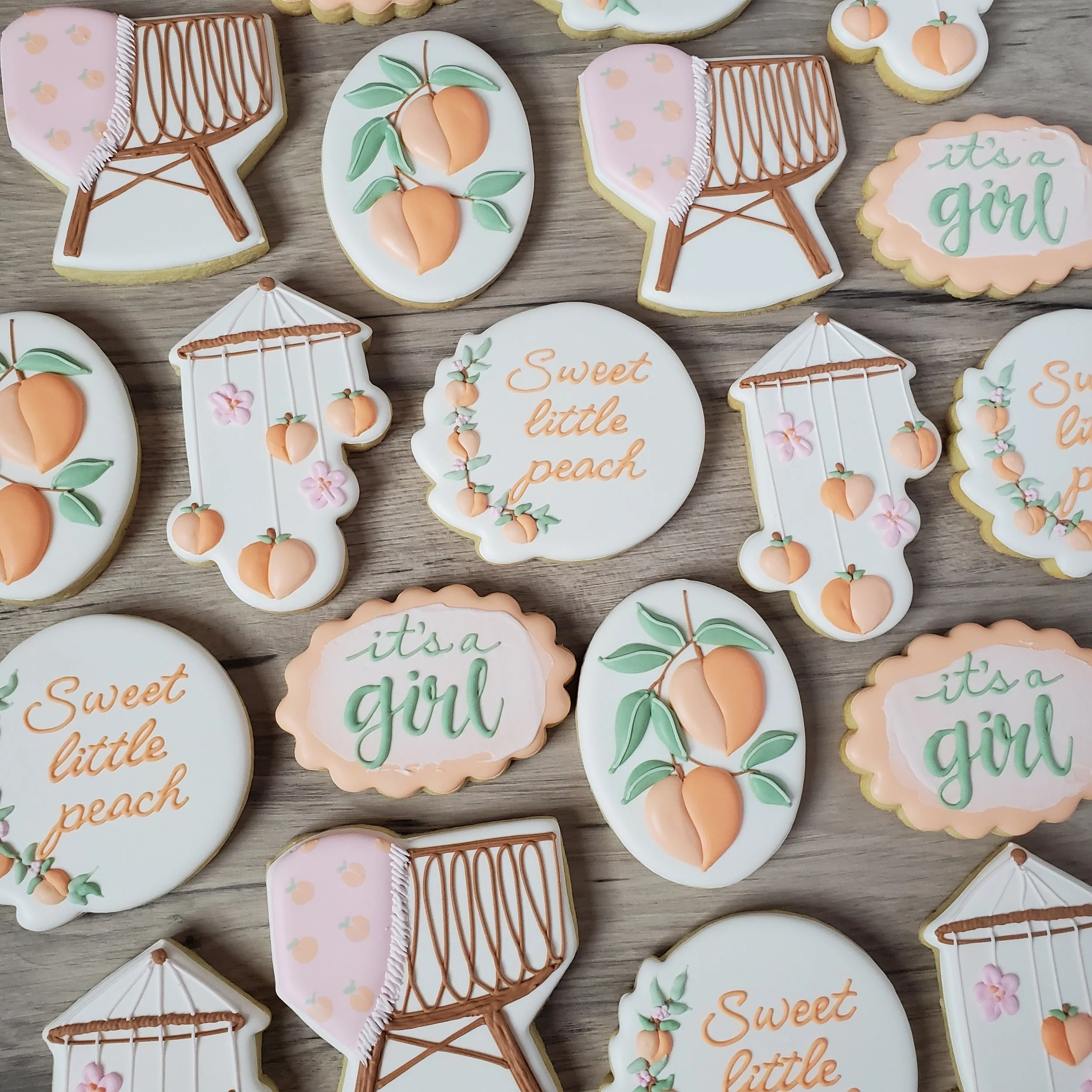 Decorated cookies with peach and peach blossom designs, and sayings like 'it's a girl' and 'sweet little peach' in pastel colors, arranged on a wooden surface.