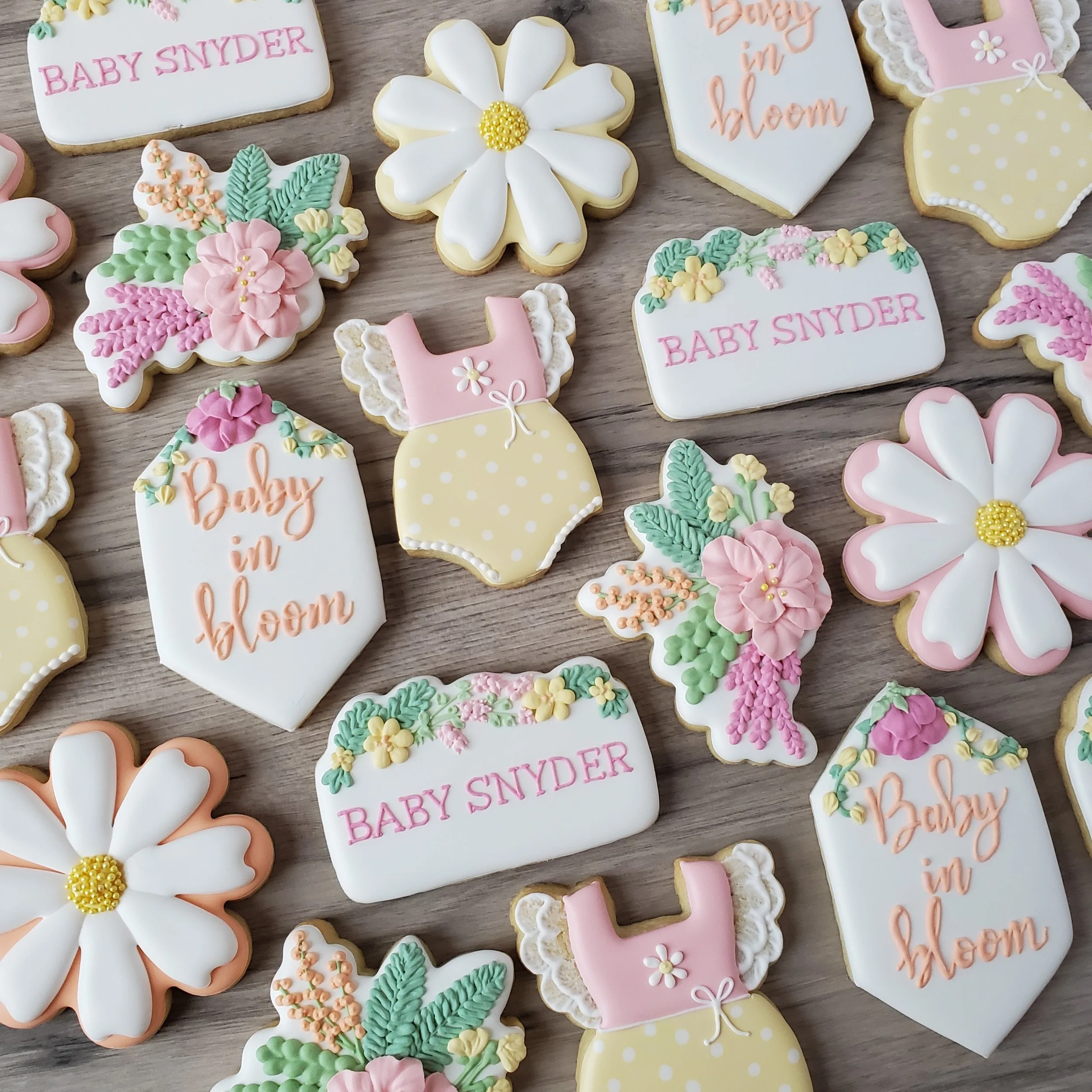 Decorated sugar cookies with floral and baby-themed designs, including flowers, baby bodysuits, and signs with phrases such as 'Baby Snyder' and 'Baby in bloom', arranged on a wooden surface.