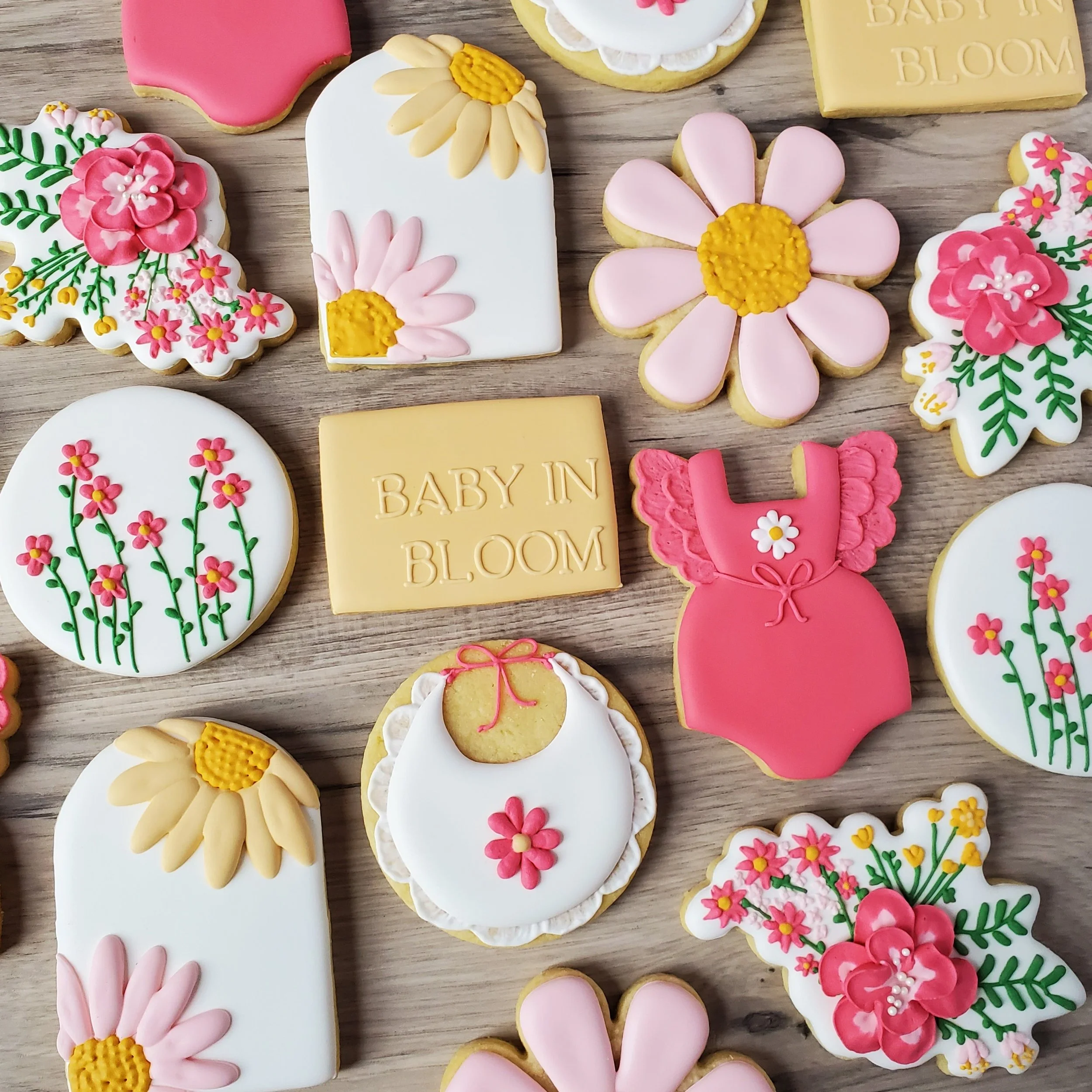 Decorative cookies with floral and baby-themed designs, including pink flowers, yellow daisies, a pink dress, a baby bib, and chocolate blocks with "BABY IN BLOOM" inscribed, arranged on a wooden surface.