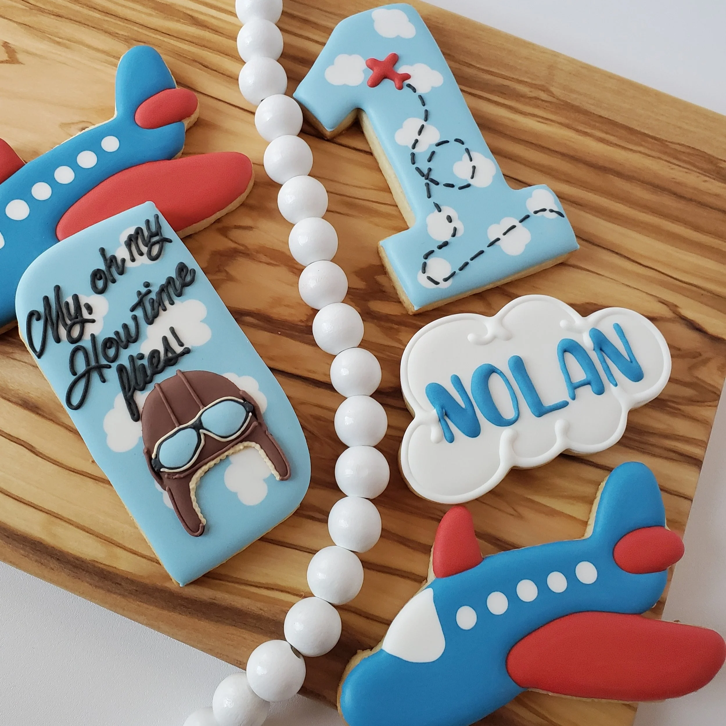 Decorative cookies with airplane and travel theme, including cookies shaped like airplanes, a number one with clouds and a red airplane, a cloud with the name Nolan, and a cookie with sunglasses and the phrase 'My oh my, Howtime flies!'. There is als