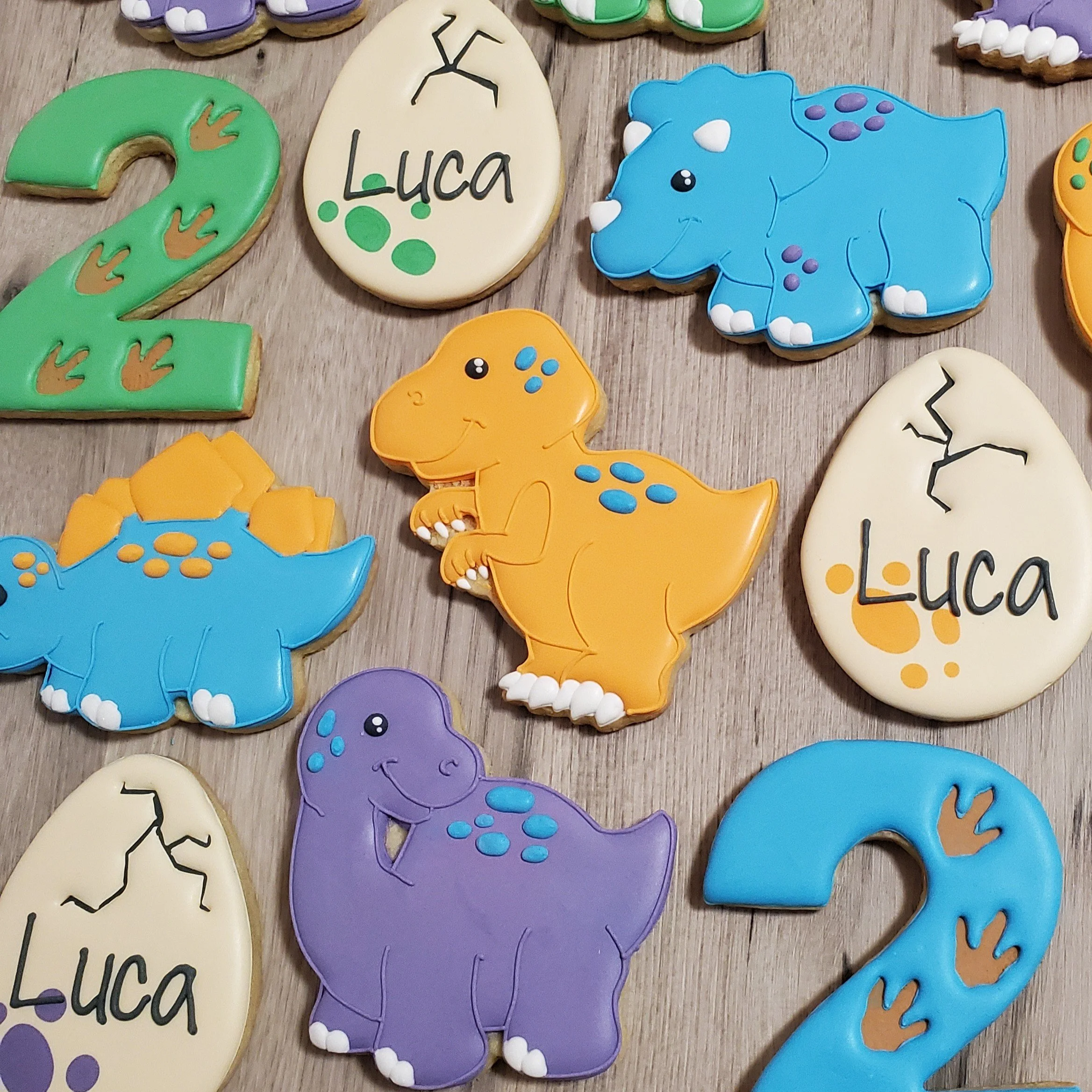Colorful dinosaur-shaped cookies and number 2 cookies decorated with fondant. Some cookies are decorated with the name 'Luca' and dinosaur footprints.