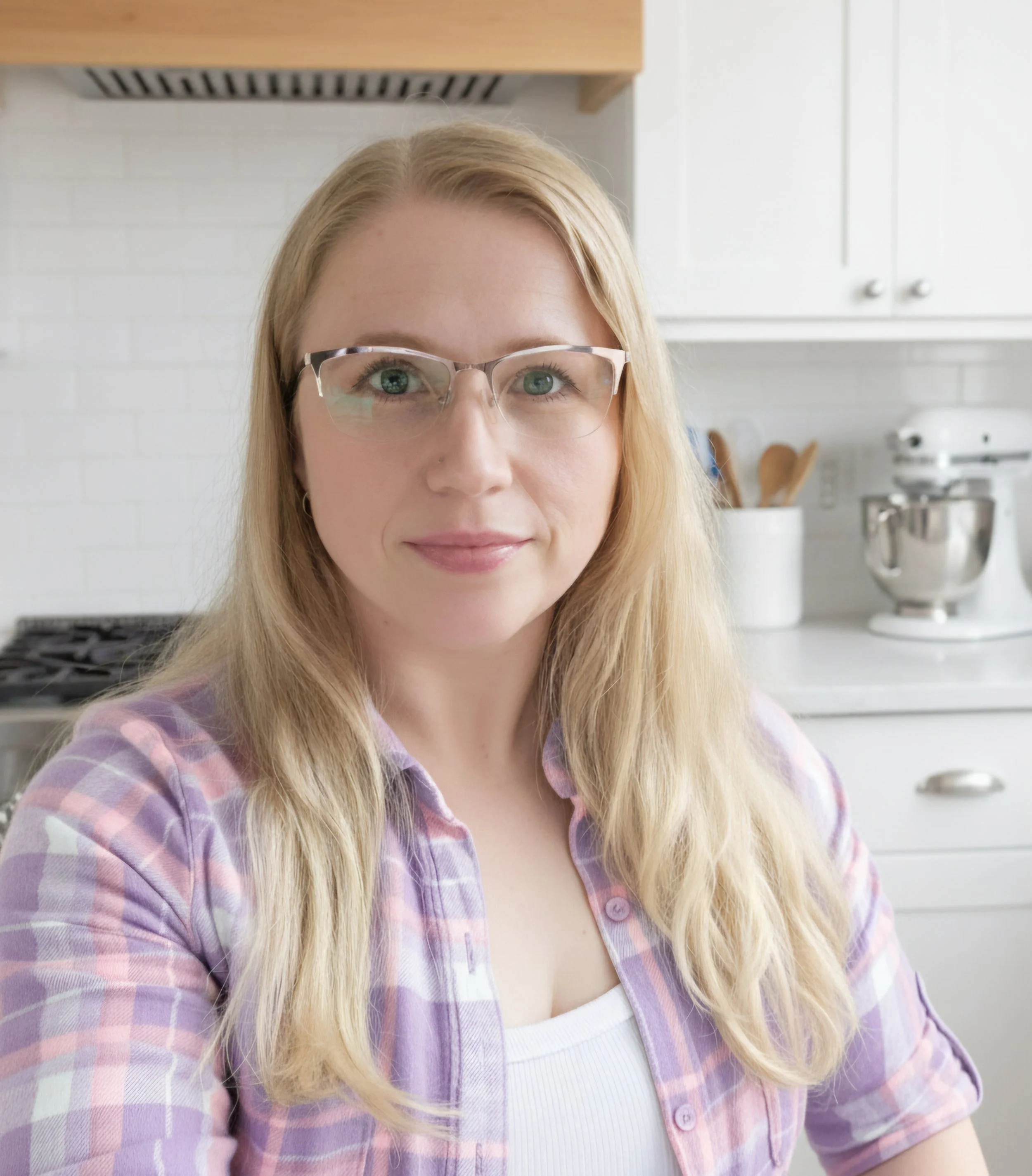 A woman with blonde hair, glasses, and a light purple plaid shirt sitting in a white kitchen, looking at the camera.