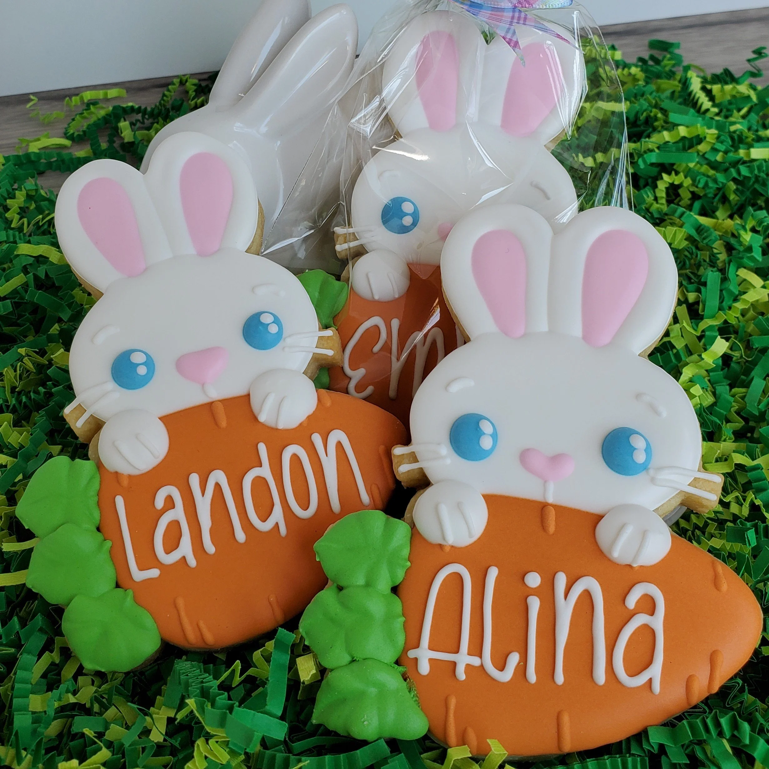 Decorated cookies with bunny faces holding orange carrots with green leaves, personalized with the names 'Landon' and 'Alina', on a bed of green and yellow decorative paper.