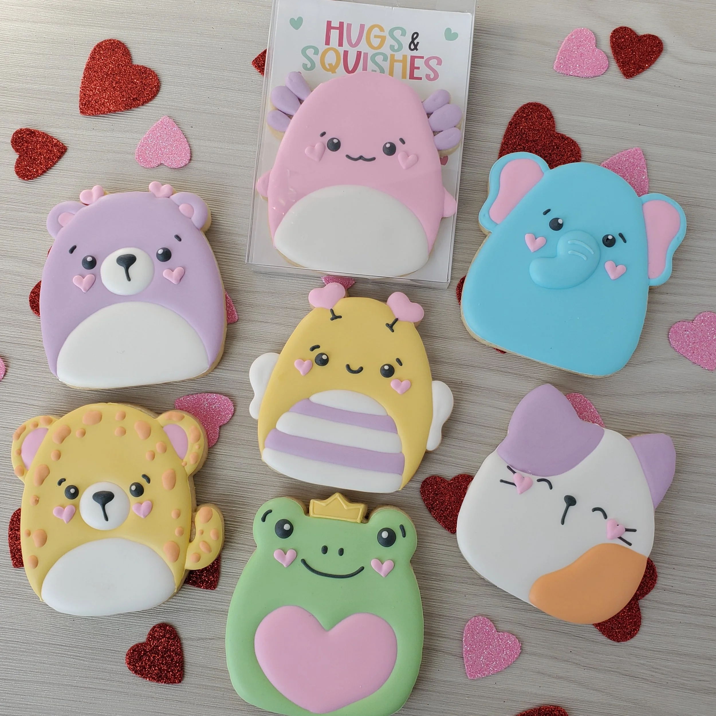 Assorted animal-shaped cookies decorated with pastel-colored icing, surrounded by red and pink glittery hearts on a light wooden surface. The cookies depict a bear, a hedgehog, a frog, an elephant, a cat, a lion, and a giraffe, all with cute facial e