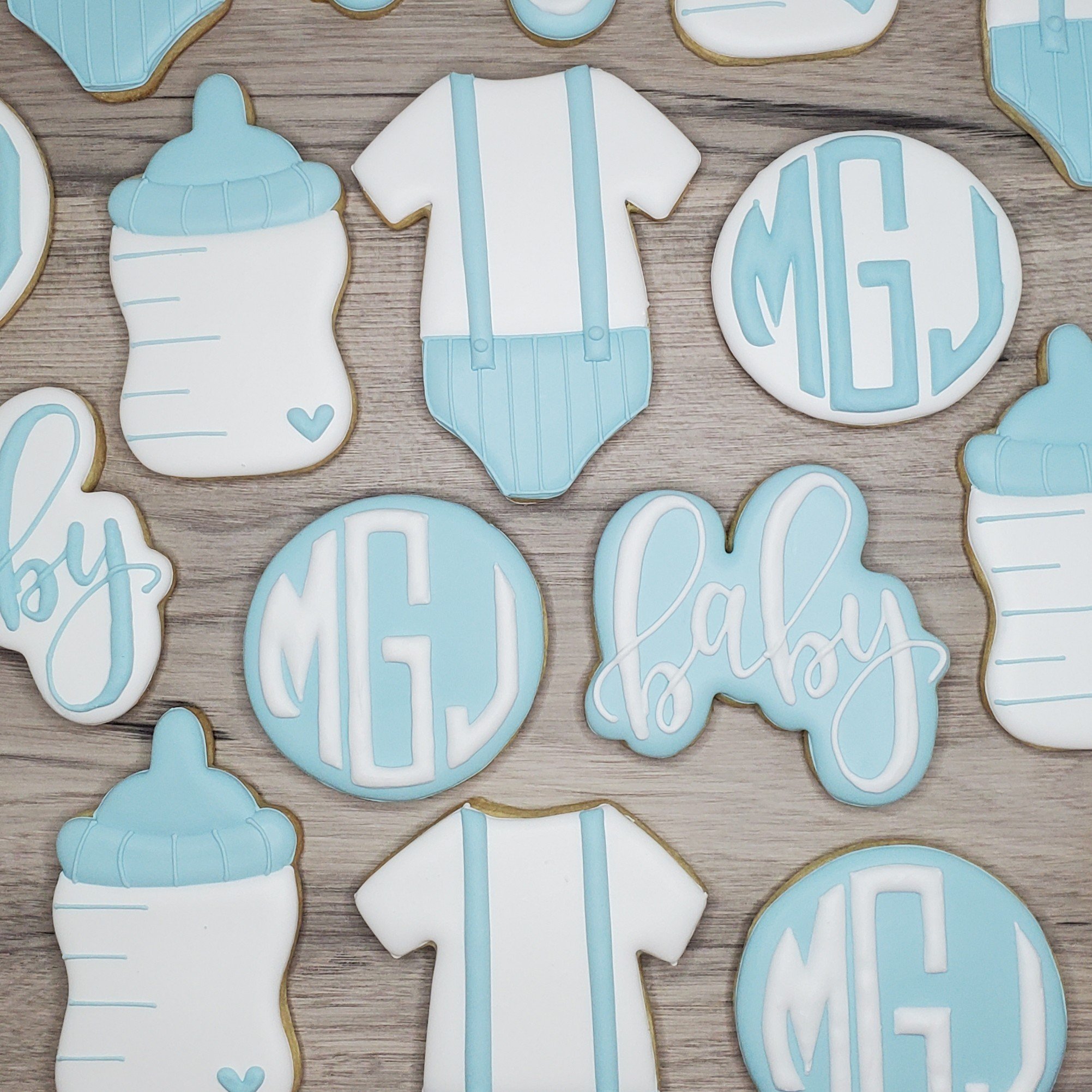 Baby shower cookies decorated with blue and white icing, including a baby bottle, a baby onesie, a round cookie with initials 'MGJ', and a cloud-shaped cookie with the word 'baby' in cursive.