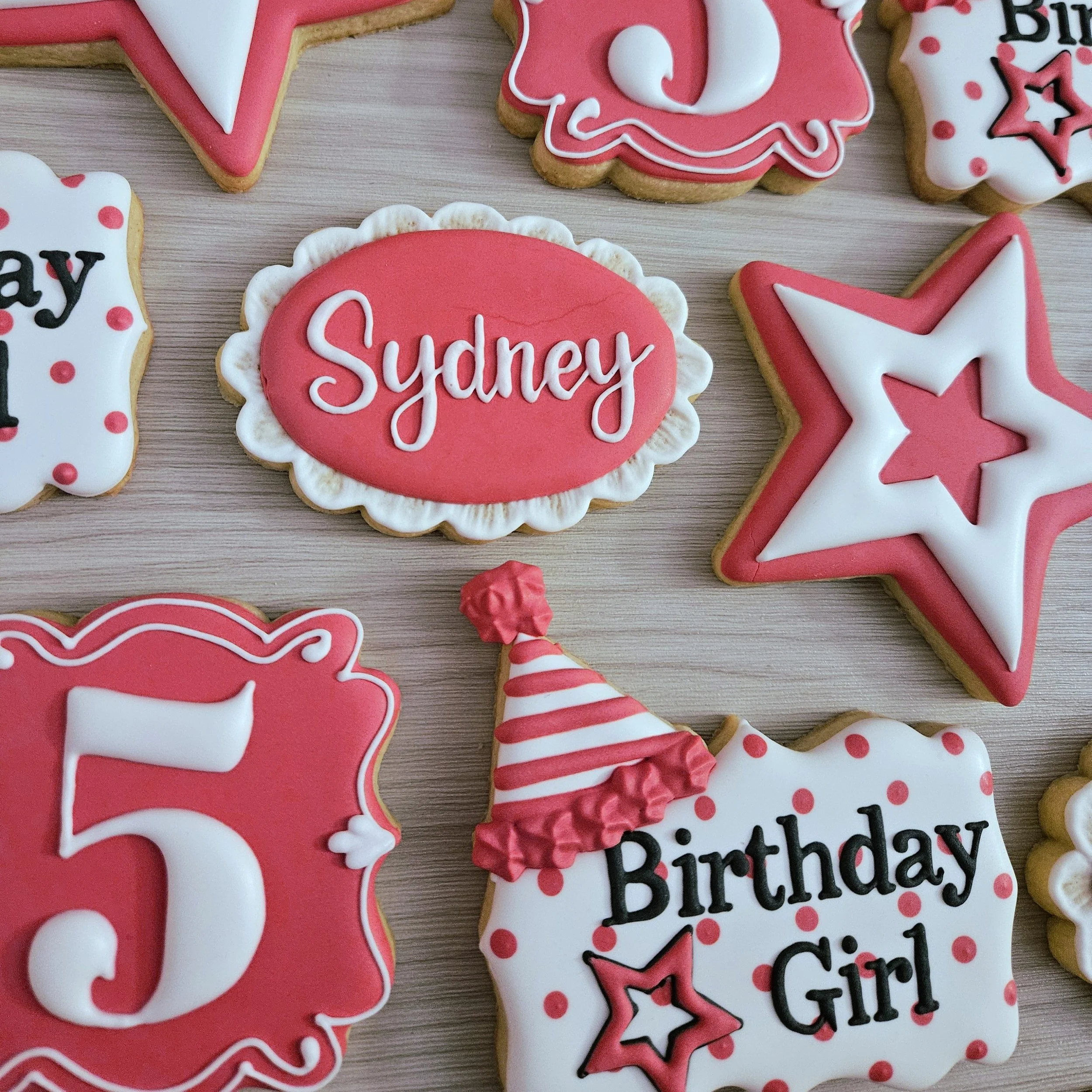 Decorated cookies for a birthday celebration, including a pink oval cookie with the name 'Sydney' in white icing, a star-shaped cookie with red and white icing, and another cookie with the text 'Birthday Girl' and a party hat, all in pink, white, and
