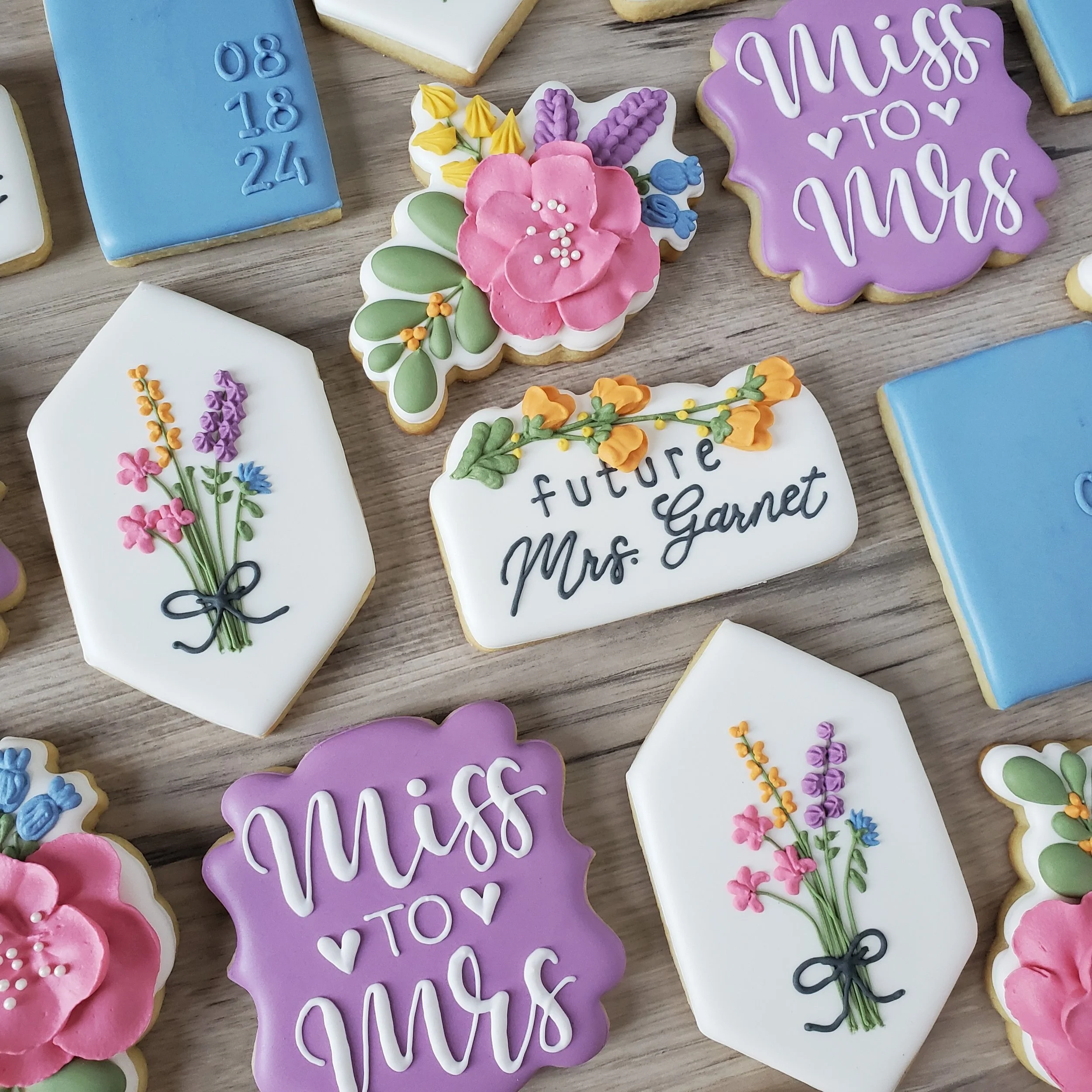 Assorted decorated cookies for a bridal shower, with messages like "Miss to Mrs" and "future Mrs. Garnet," and floral designs in bright colors on a wooden surface.