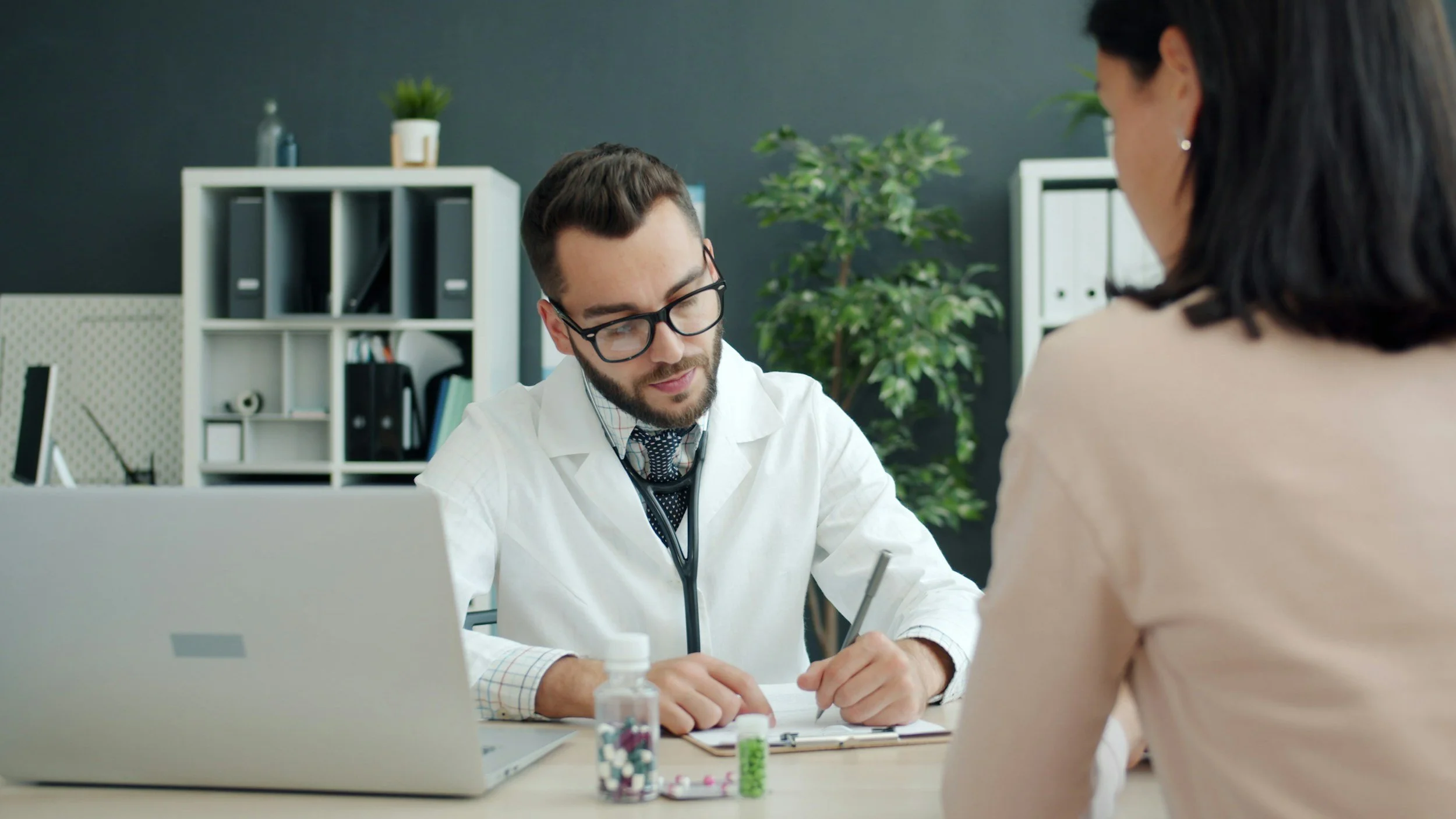 A doctor in white coat and glasses consulting a woman at a desk, with medication bottles in front and a laptop behind.