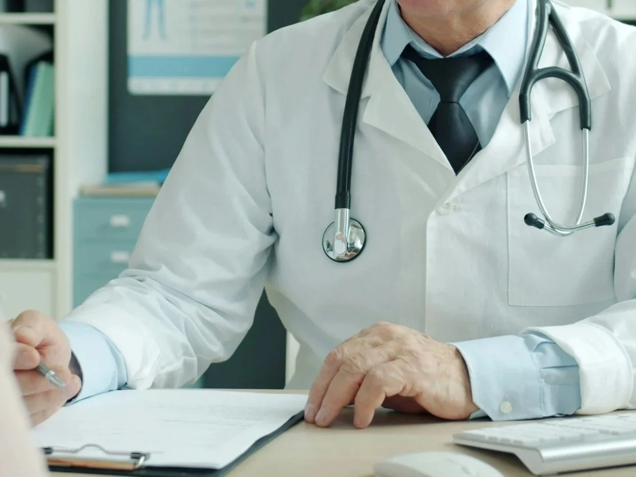 A doctor in a white coat with a stethoscope around his neck taking notes at a desk in an office