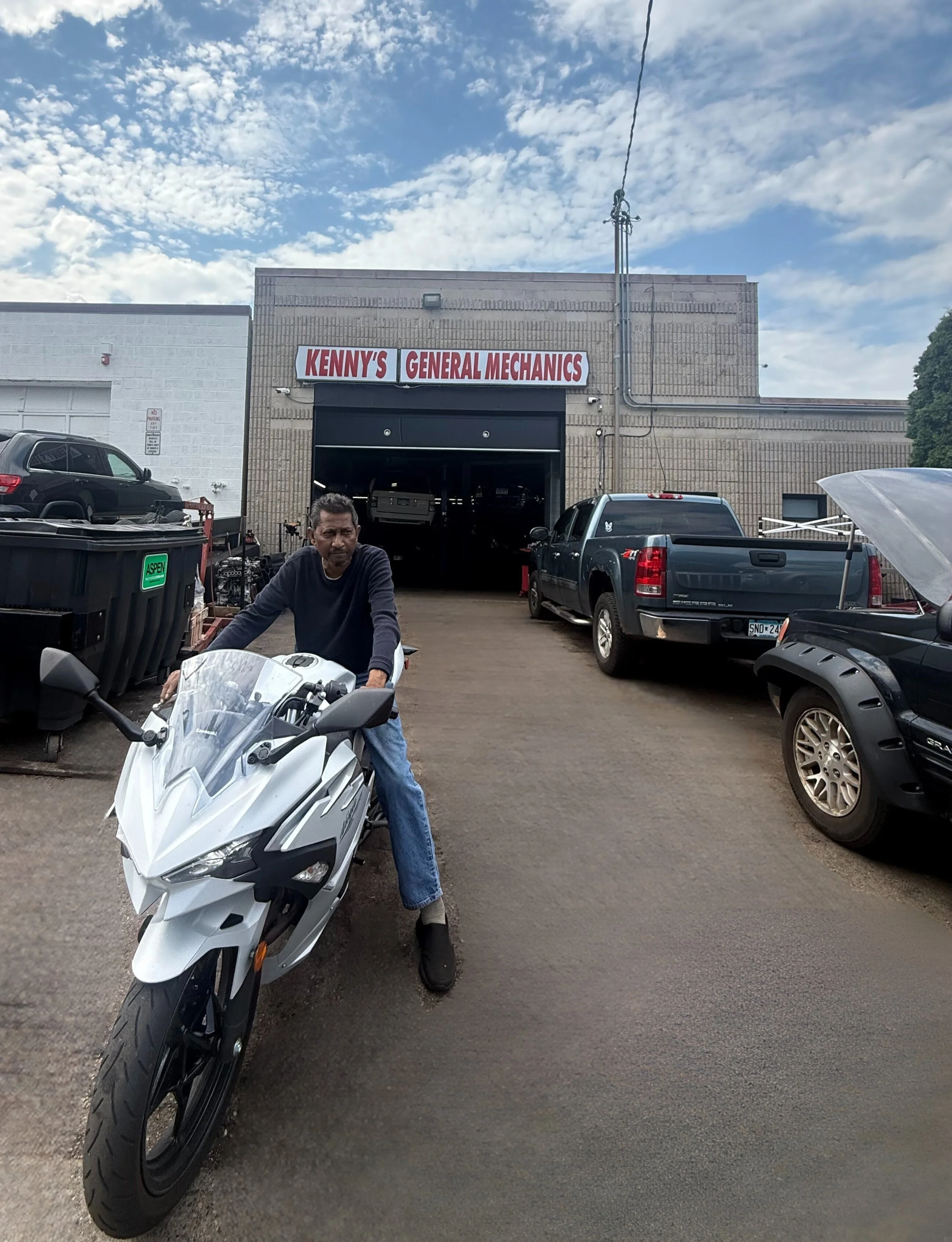 A man sitting on a white motorcycle in front of a garage with a sign that reads 'Kenny's General Mechanics,' several parked vehicles, and a bright partly cloudy sky.