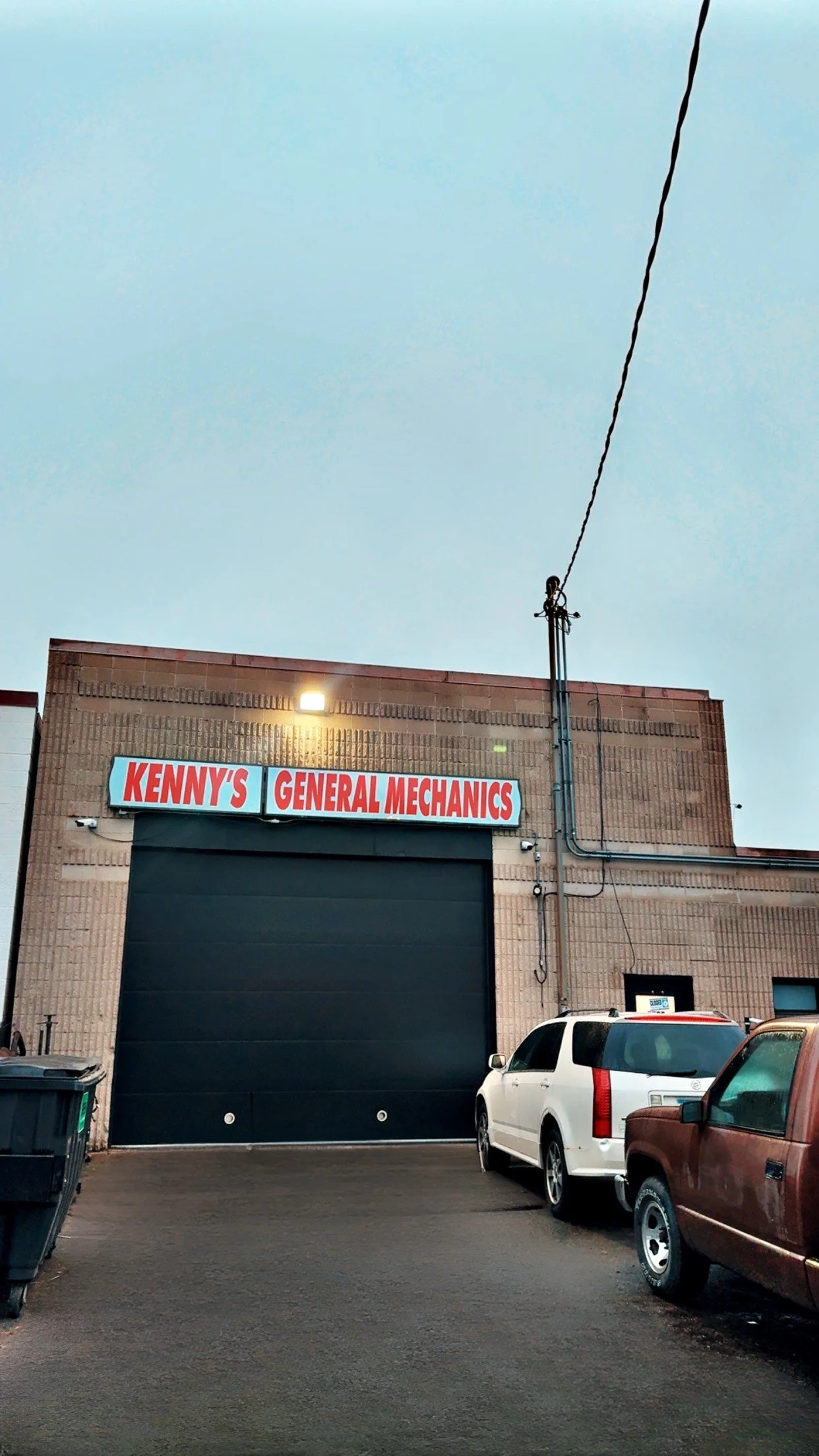 Exterior of an auto repair shop named Kenny's General Mechanics with a large black garage door, parked cars, and electrical wires on the building