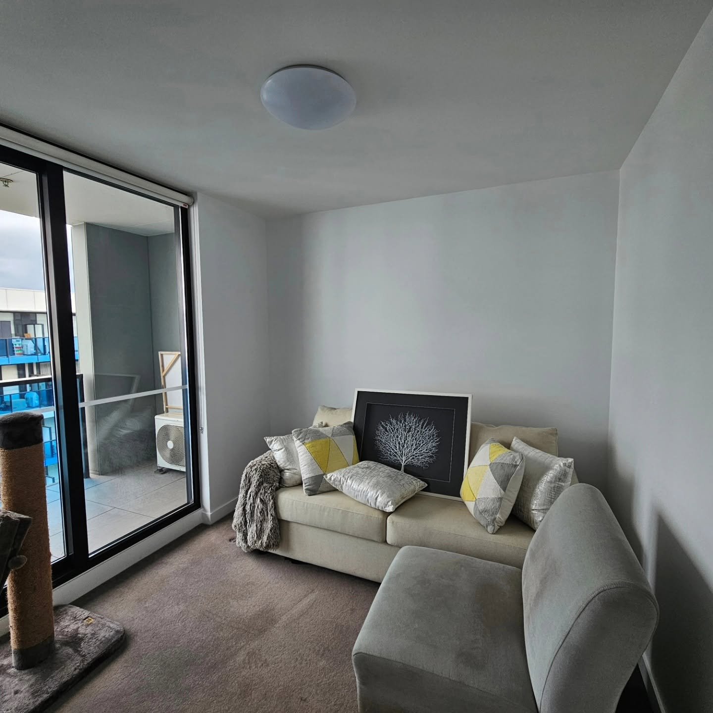 Living room with beige sofa, decorative cushions, artwork of a tree, sliding glass door leading to a balcony with outdoor air conditioning unit, beige carpet, and plain white walls.
