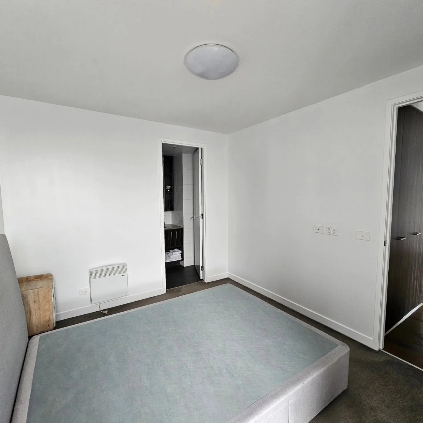 Empty room with white walls and a bed with light blue mattress, doorway leading to a bathroom with stacked towels, dark wood shelves, and a small bathroom heater, and a doorway to another room with dark wood door.