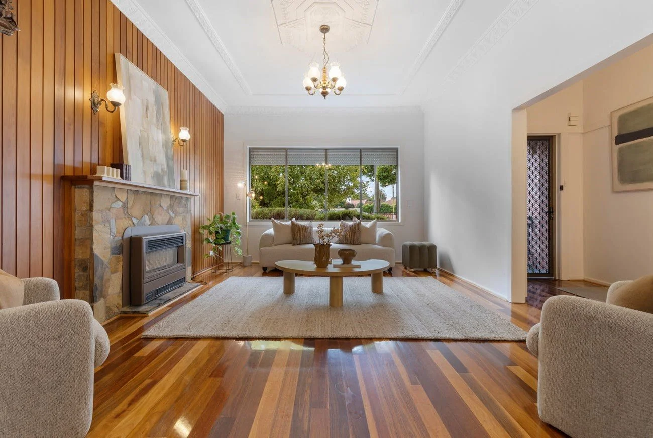 Living room with wooden floor, a cream sofa, and two matching armchairs. A stone fireplace with a wooden mantel and decorative items. A large window with greenery outside. Ceiling light fixture, beige area rug, and modern wall art.