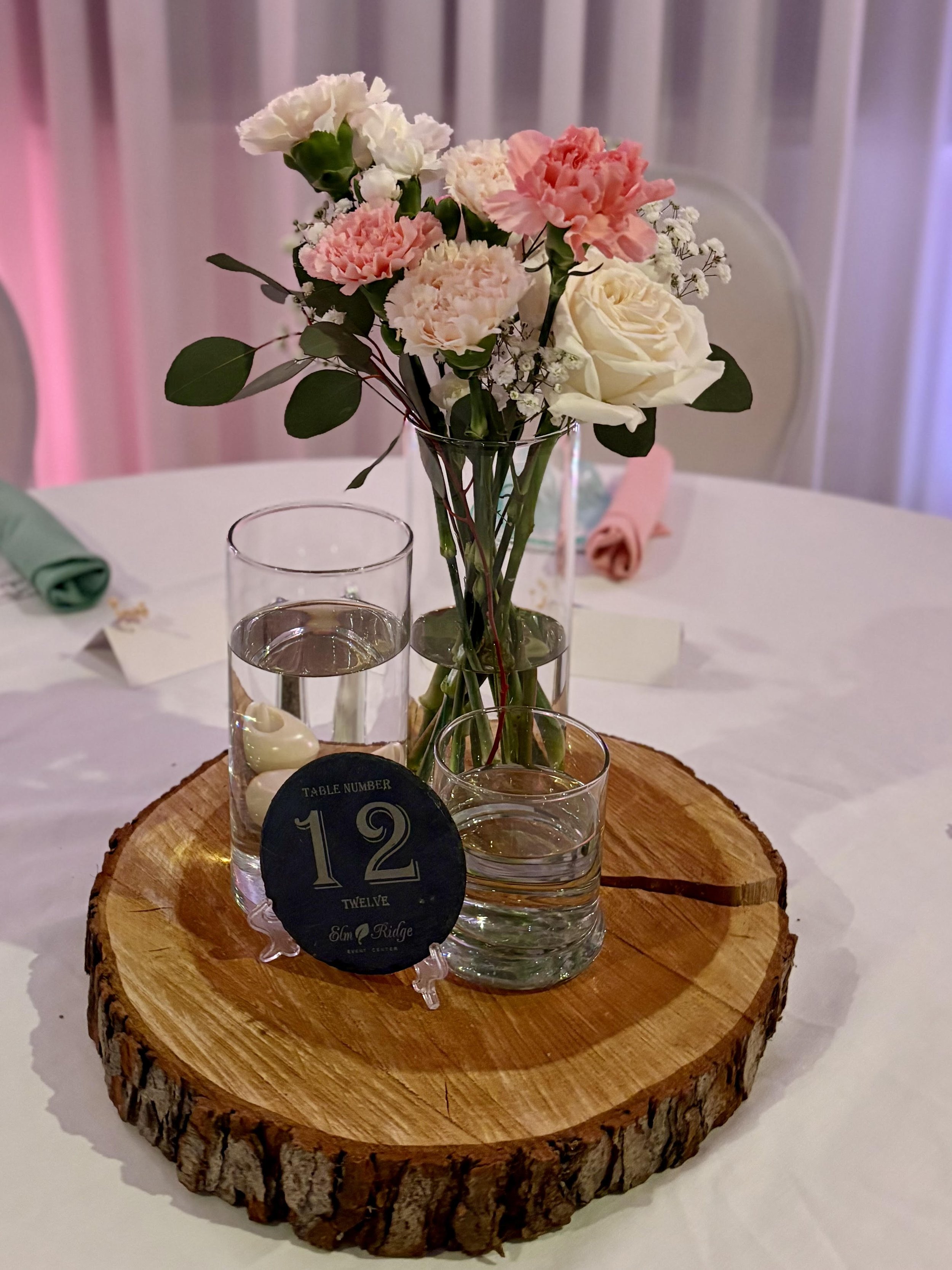 A centerpiece on a table featuring a glass vase with pink and white flowers, three glasses with water, and a table number card on a round wooden slice.