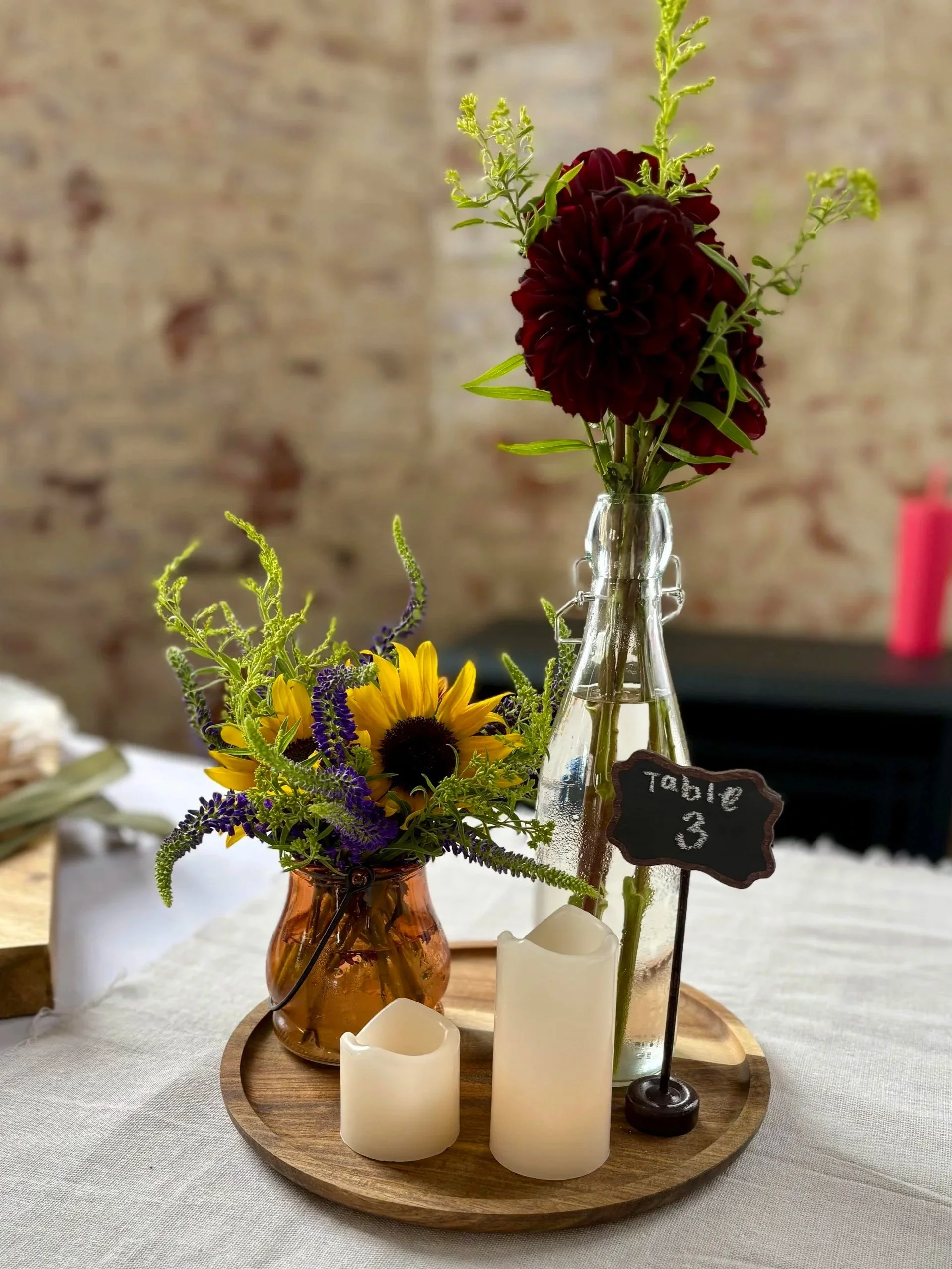 Decorative table centerpiece with two vases holding flowers and candles, and a small chalkboard sign labeled 'table 3' on a round wooden tray.
