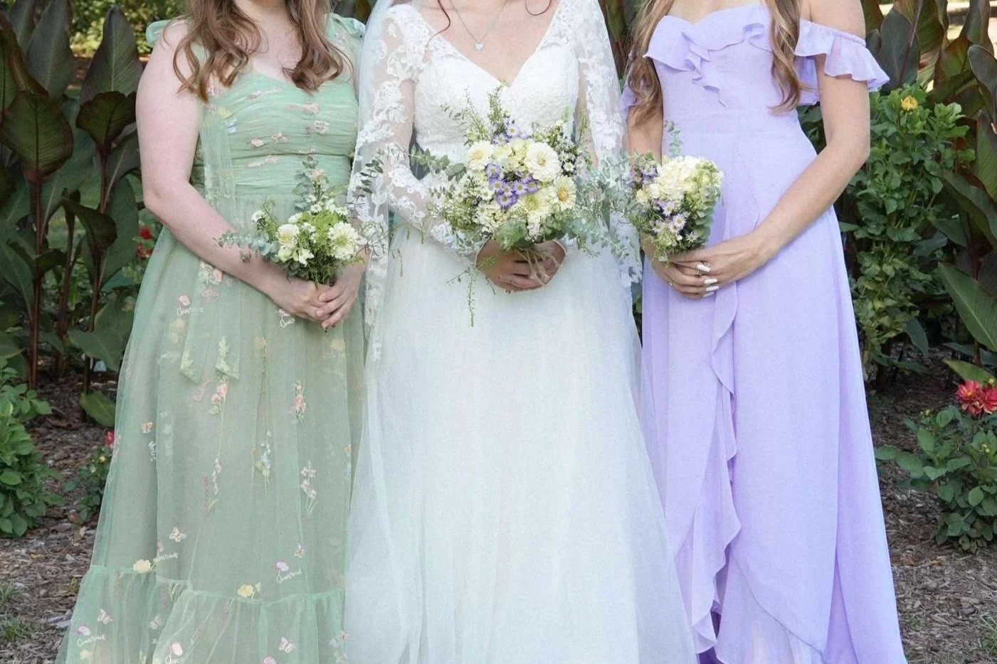 A bride standing between two women, each holding a bouquet. The bride wears a long white lace dress, and the women wear pastel-colored dresses, light green and lavender, all holding small floral arrangements. They are outdoors surrounded by green plants and flowers.
